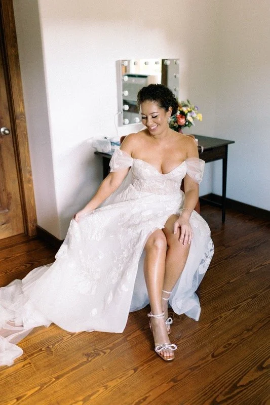 A woman in a wedding dress sitting on a chair, smiling, with one leg crossed over the other, in a room with wooden floors and a mirror.