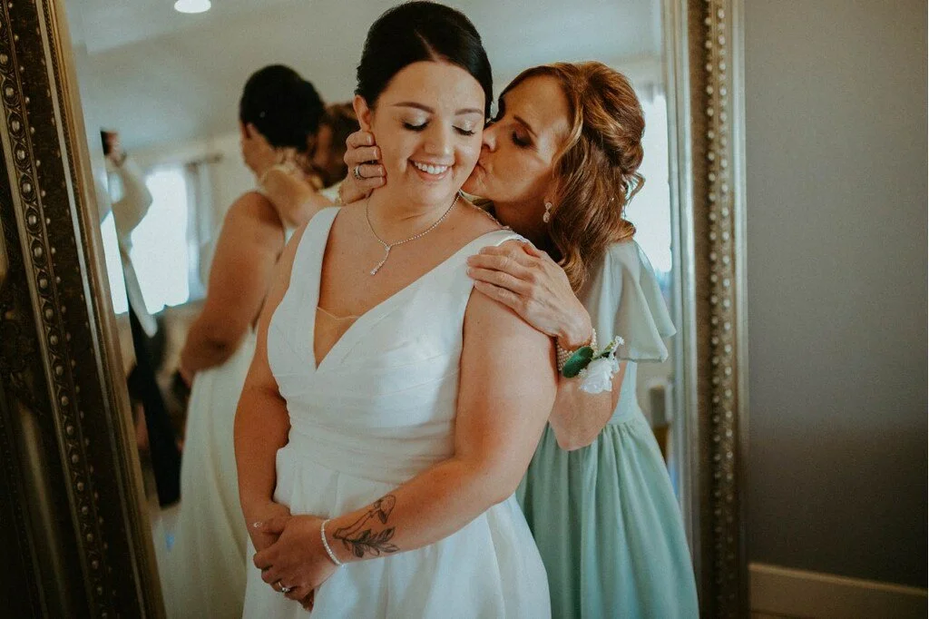 A woman in a white dress has her eyes closed and is smiling while an older woman kisses her on the cheek. They are standing in front of a mirror, dressed in formal attire, likely at a wedding or special occasion.