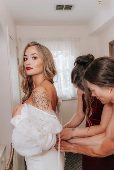 A woman with blonde hair and red lipstick standing while two other women assist her with her dress in a bright room.