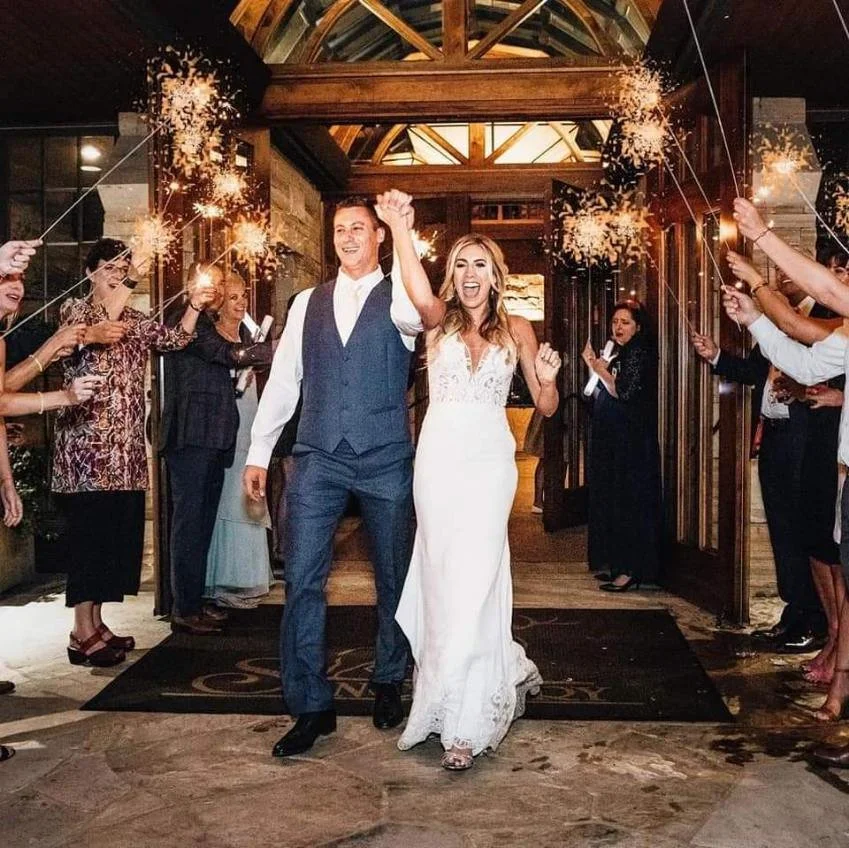 A newlywed couple walking through a sparkler exit, smiling and celebrating with friends and family at a wedding reception.