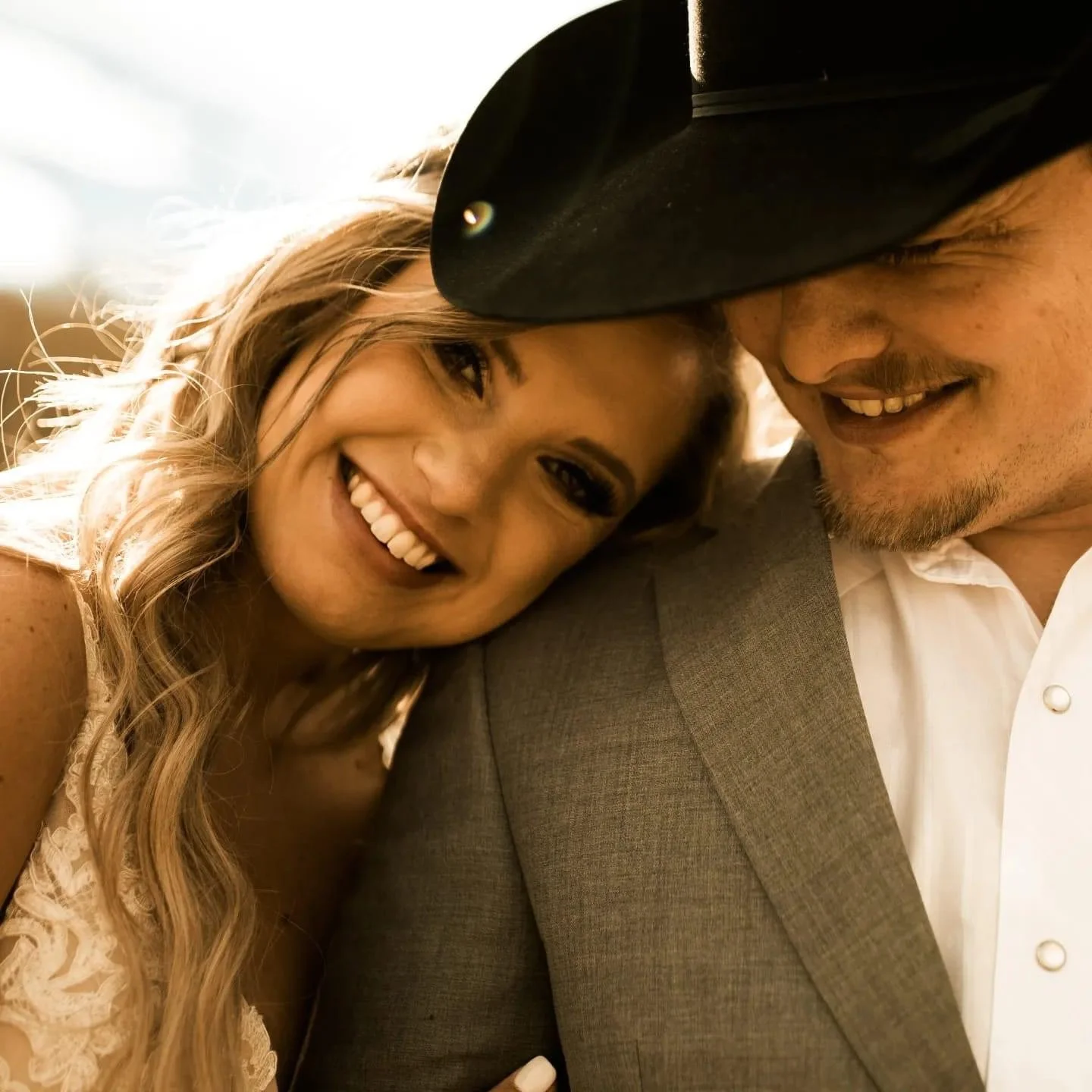 A happy woman with long, wavy hair leaning her head on a man’s shoulder. The man is wearing a black cowboy hat and a gray suit. They are smiling and close together.