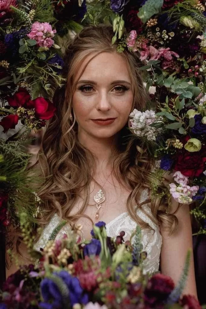 A woman with long, wavy hair surrounded by a variety of colorful flowers, wearing a white dress and a necklace, with a confident expression.