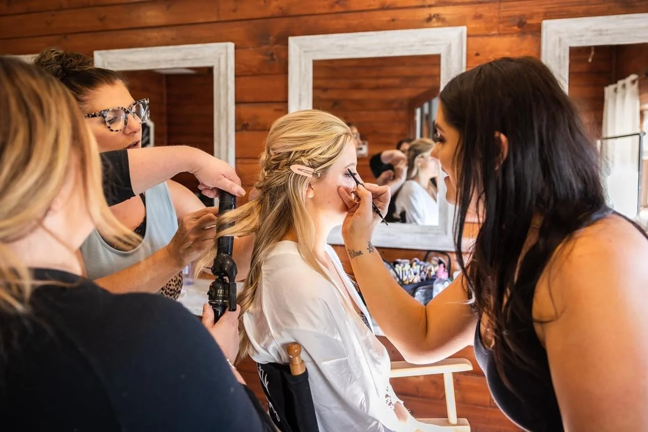 A woman is getting her makeup done by a makeup artist while her hair is being styled with a curling iron, with other women reflected in the mirror.