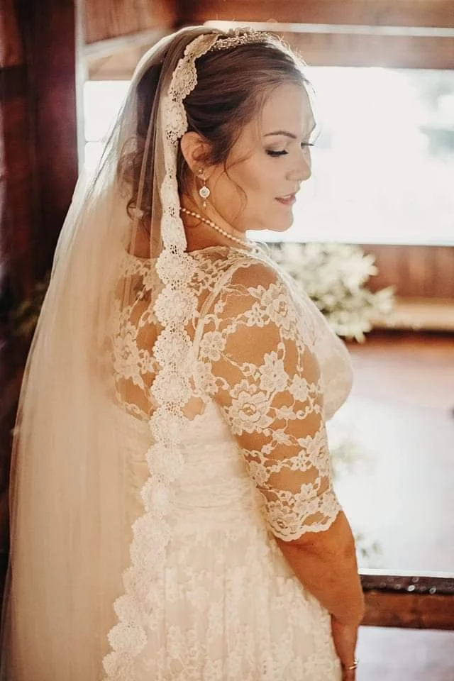 A bride in a white lace wedding dress with long sleeves, lace veil, and pearl jewelry, standing inside a wooden building with sunlight coming through a window.