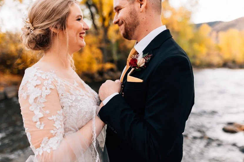 A bride and groom facing each other outdoors near a river with autumn trees in the background, holding hands and smiling.