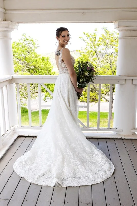 A bride in a lace wedding dress holding a bouquet, standing on a porch with a white railing, smiling and looking over her shoulder, with green trees and grass in the background.