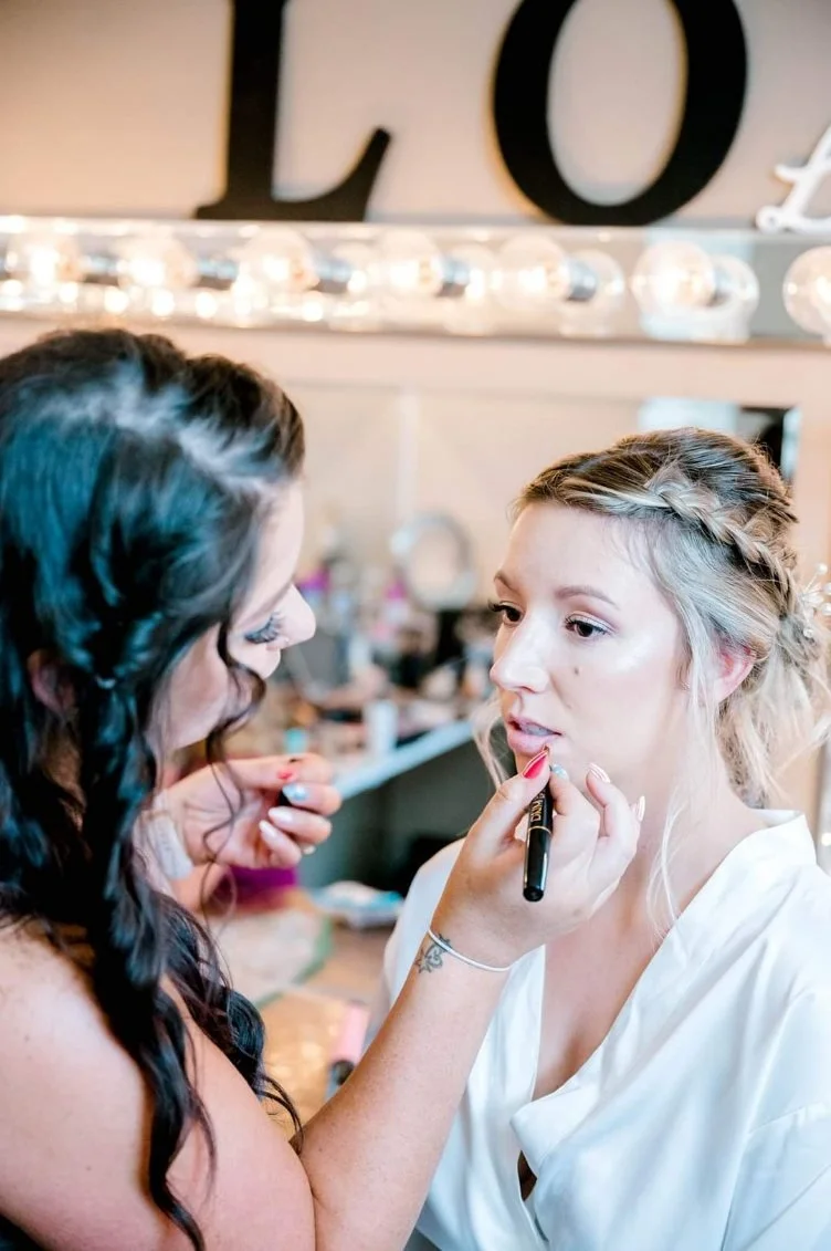 A makeup artist applying lipstick to a woman with blonde hair styled in a braid, in a room decorated with letters and lights.