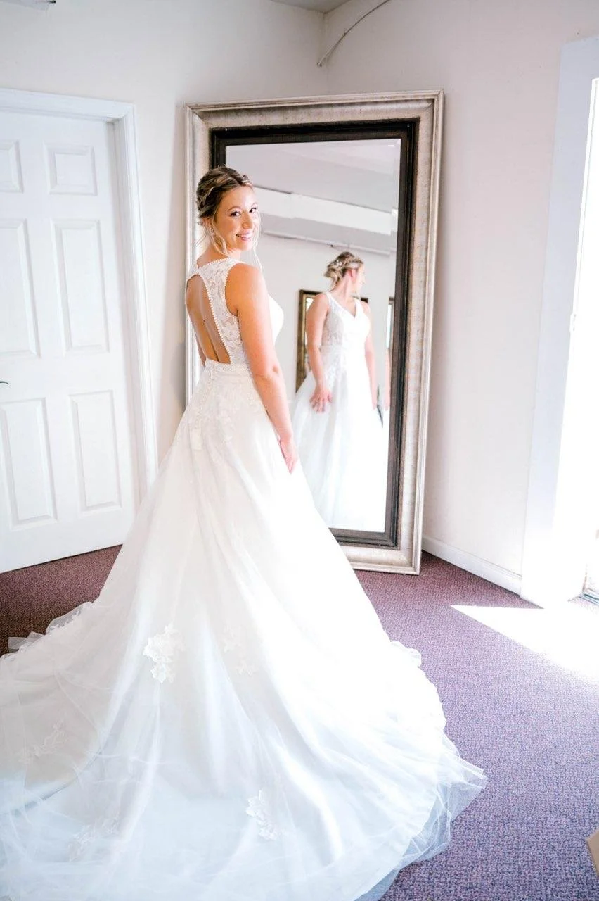 Bride in a white wedding gown standing in front of a mirror, smiling, with her reflection visible.