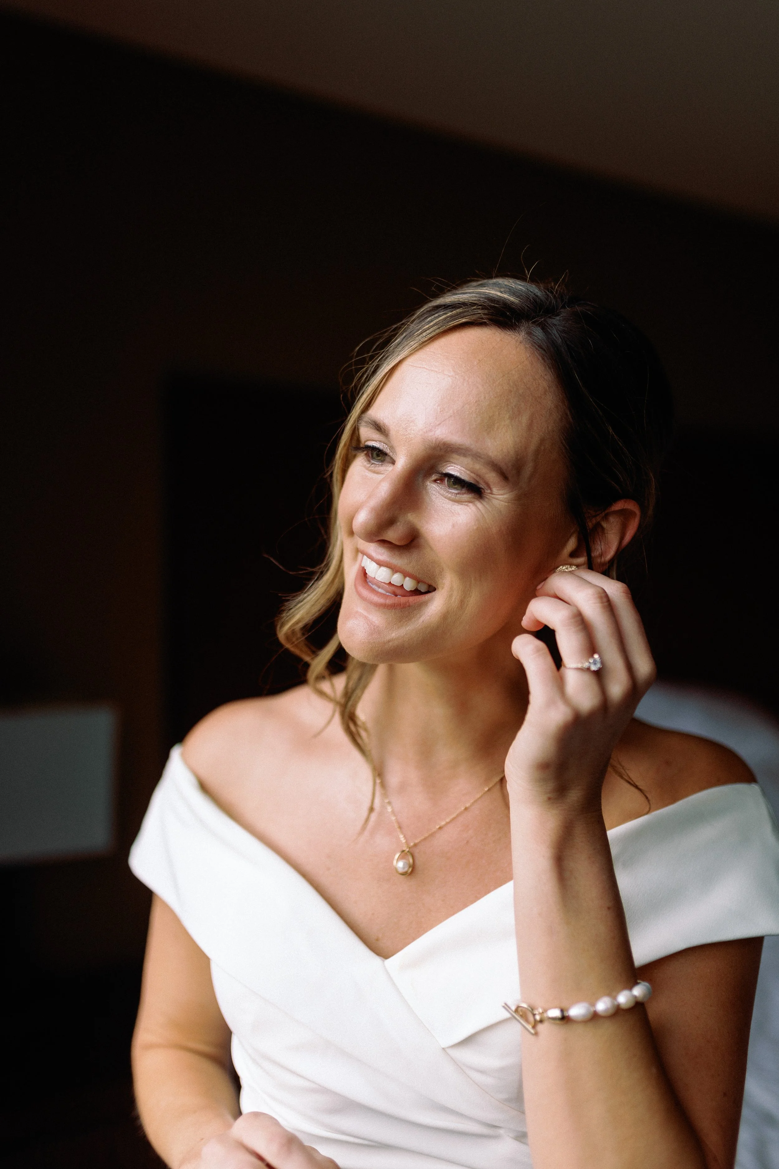 Close-up of a smiling woman with light brown hair, wearing a white off-shoulder dress, pearl jewelry, and an engagement ring, adjusting her earring indoors.