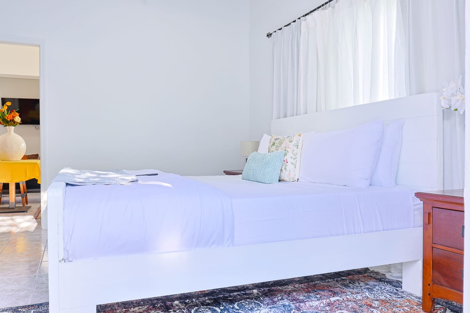 A neatly made bed with white bedding and several pillows, situated by a window with white curtains in a bright, minimalistic bedroom.