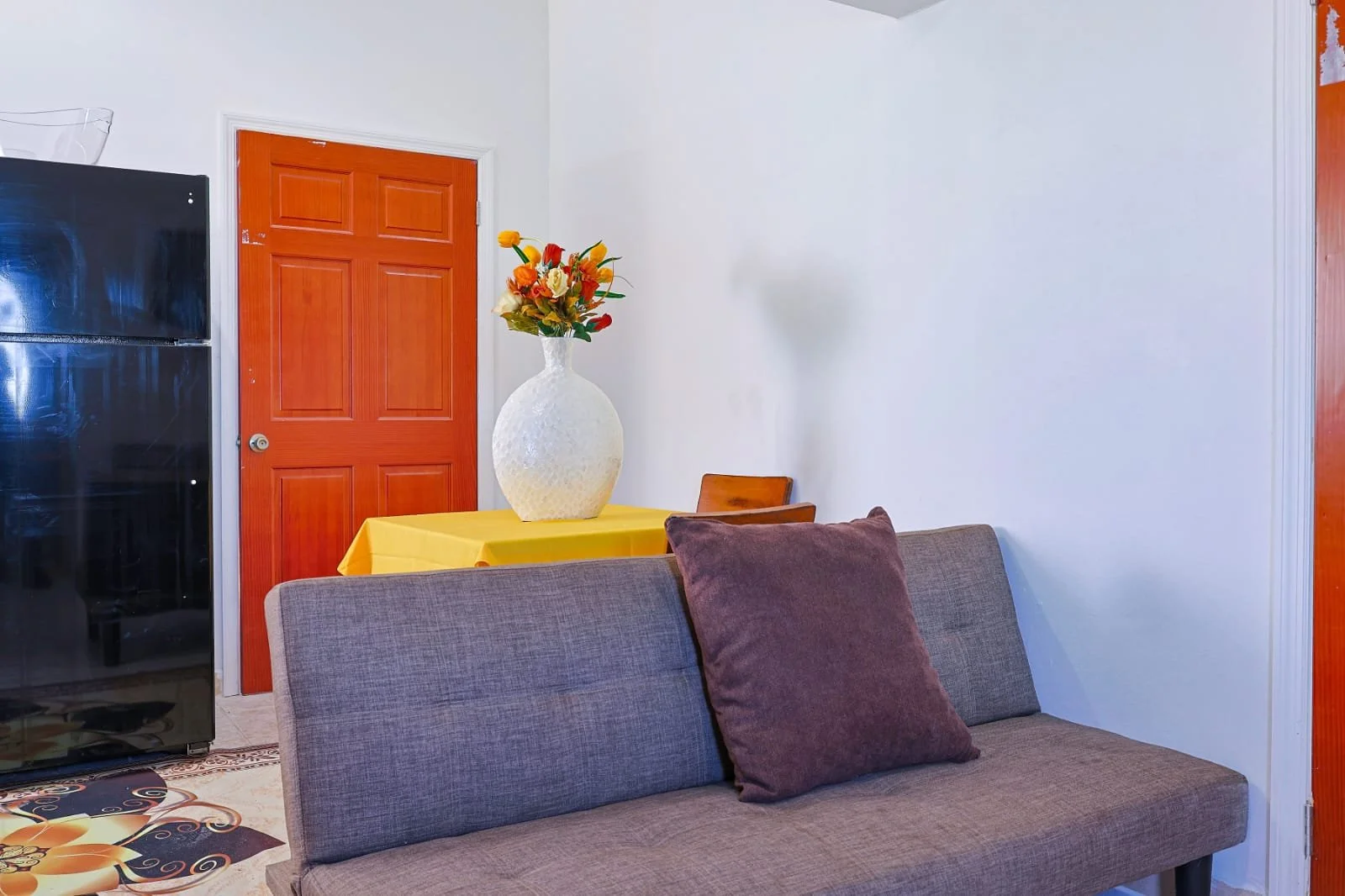 Living room with a gray sofa, brown pillows, a wooden door, a yellow tablecloth with a large white vase of colorful flowers, and a black refrigerator.