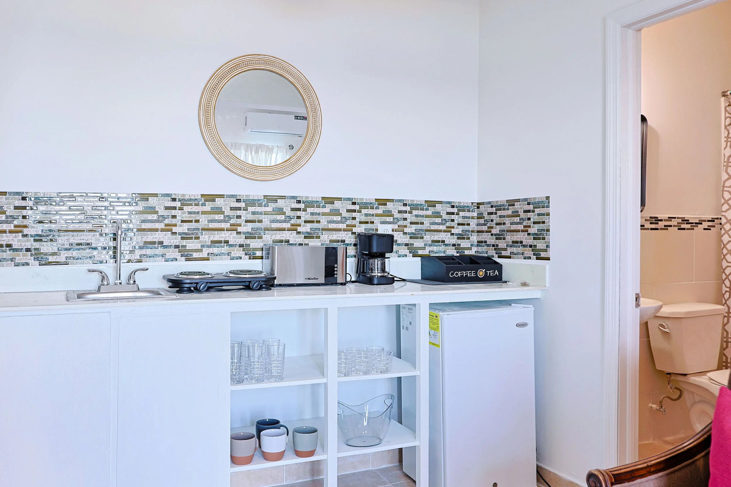Mini kitchenette with a sink, microwave, coffee maker, stove, and glassware, with a partial view of a restroom with a toilet in the background.
