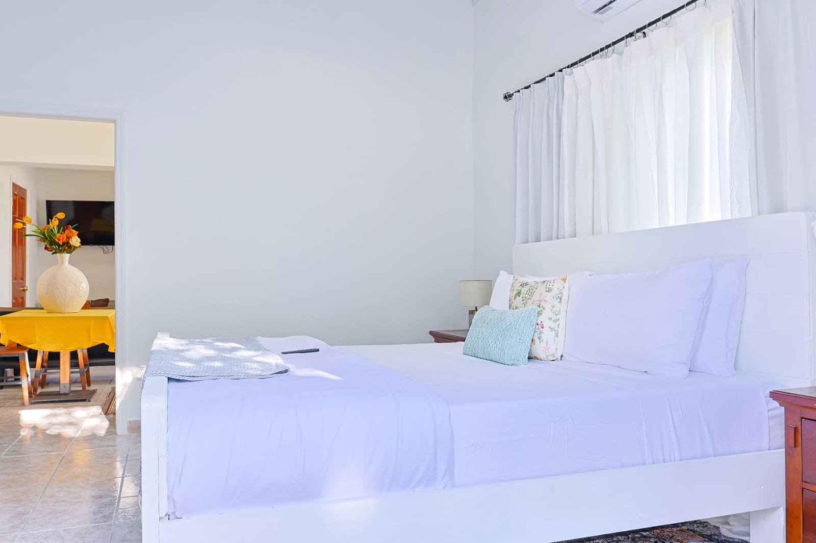 A bright bedroom with white bedding, pillows, and curtains, with a wooden nightstand and a view into another room with a yellow tablecloth, vase with flowers, and a mounted TV.