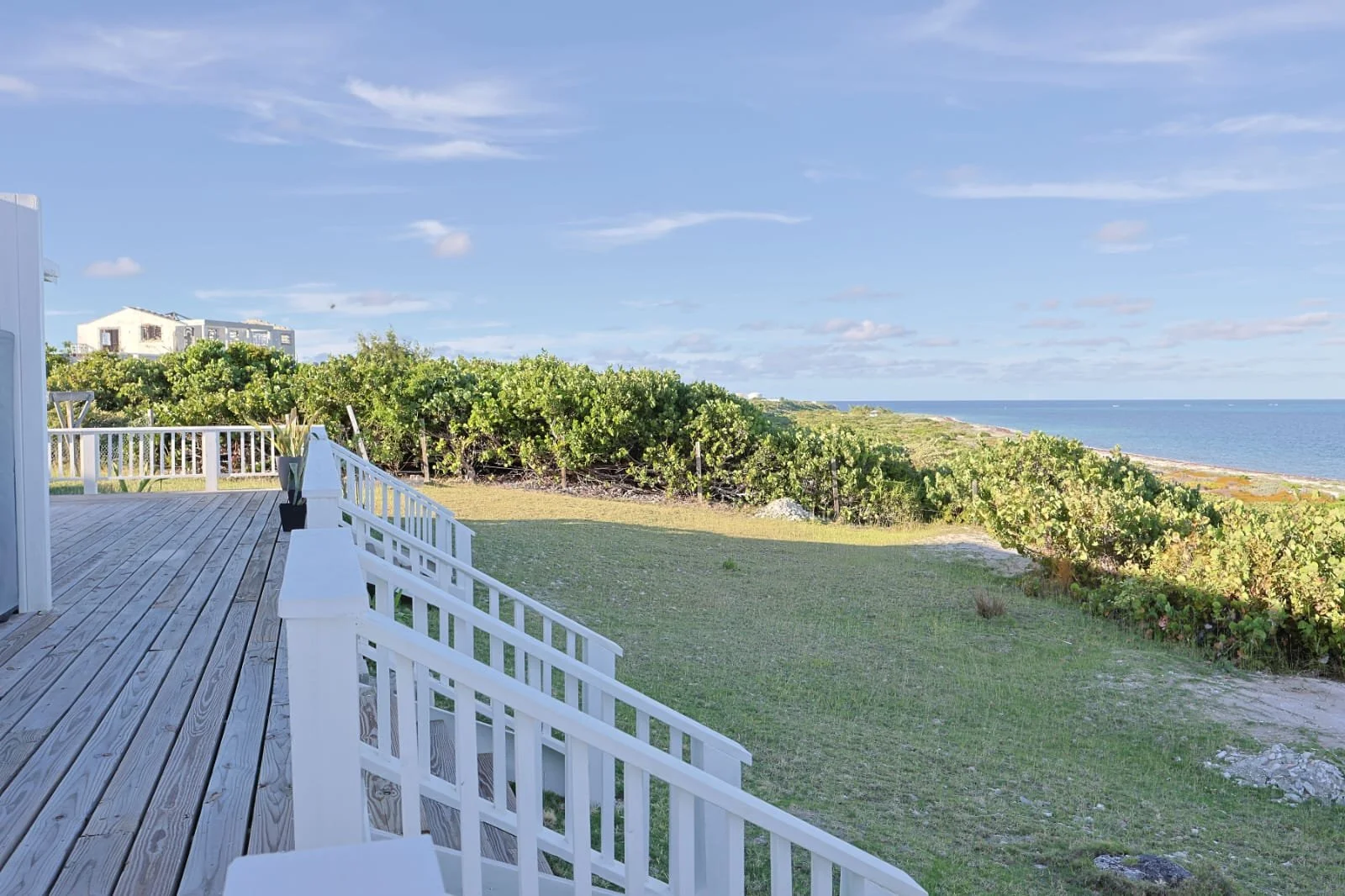 Beachside deck with white railing overlooking green shrubs, sandy beach, and ocean under a blue sky with scattered clouds.