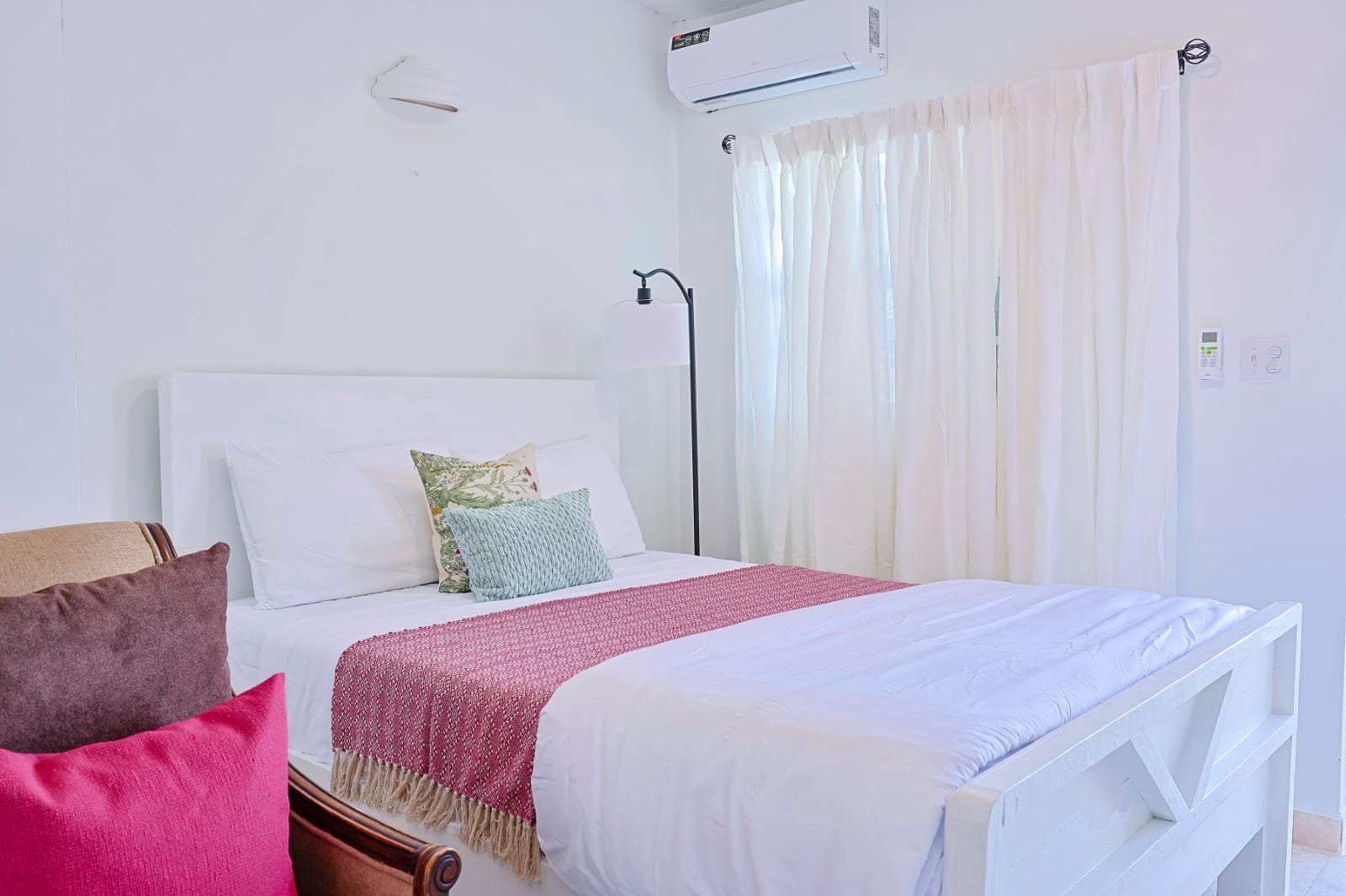A bedroom with a white bed, colorful pillows, a floor lamp, sheer curtains, and an air conditioning unit on the wall.