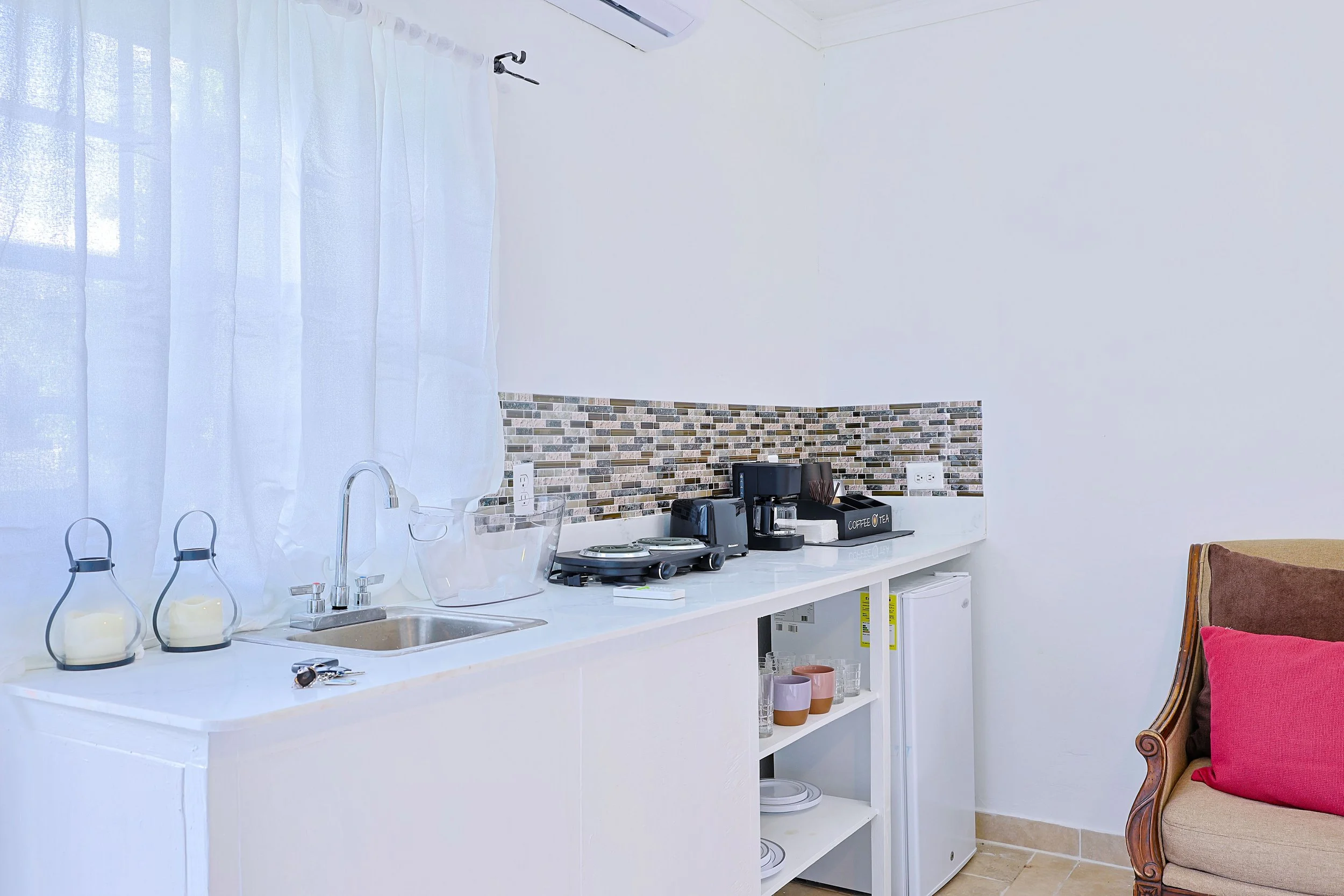 Kitchenette with white countertop and backsplash tiles, coffee maker, toaster, glassware, and a sink, adjacent to a beige armchair with a pink pillow.