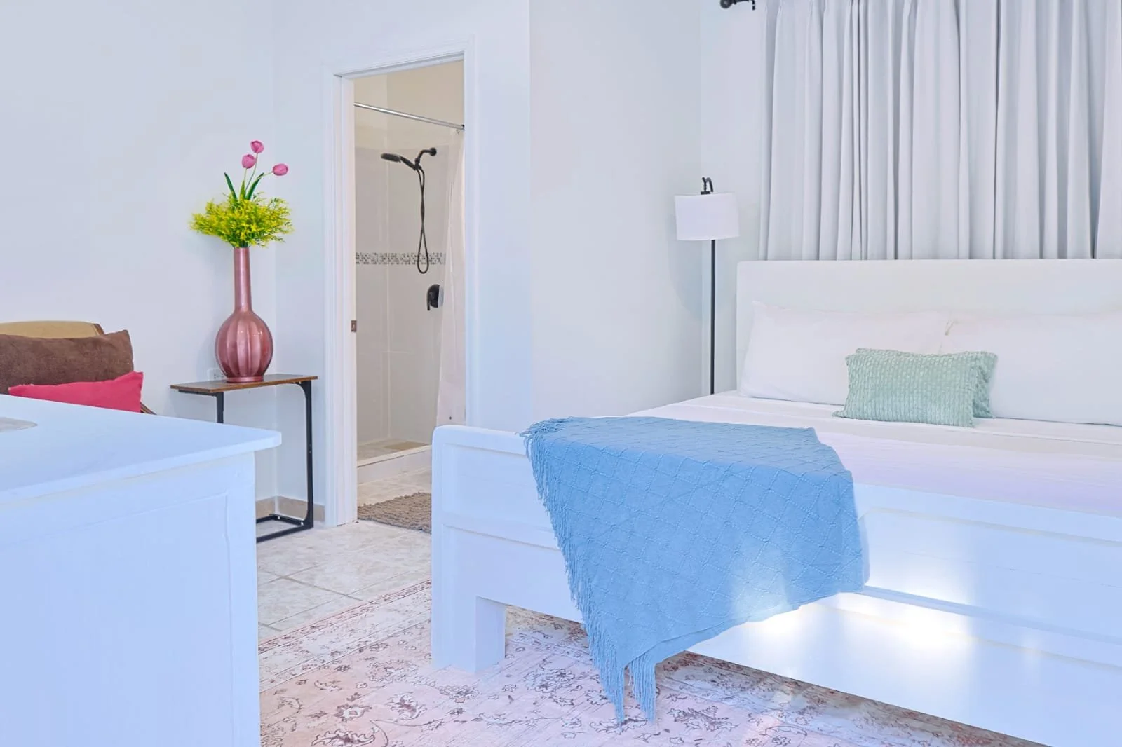 A bright bedroom with white walls, a neatly made bed with white linens and a mint green pillow, a blue throw blanket, a white floor lamp, and a window with white curtains. There is a door leading to a bathroom with a shower visible.