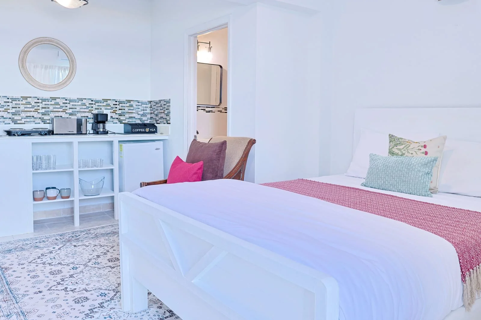 A bedroom with a white bed, pink and light blue pillows, a wooden armchair with a pink pillow, and a kitchenette area with a mini fridge, microwave, coffee maker, and a tray with cups and glasses.