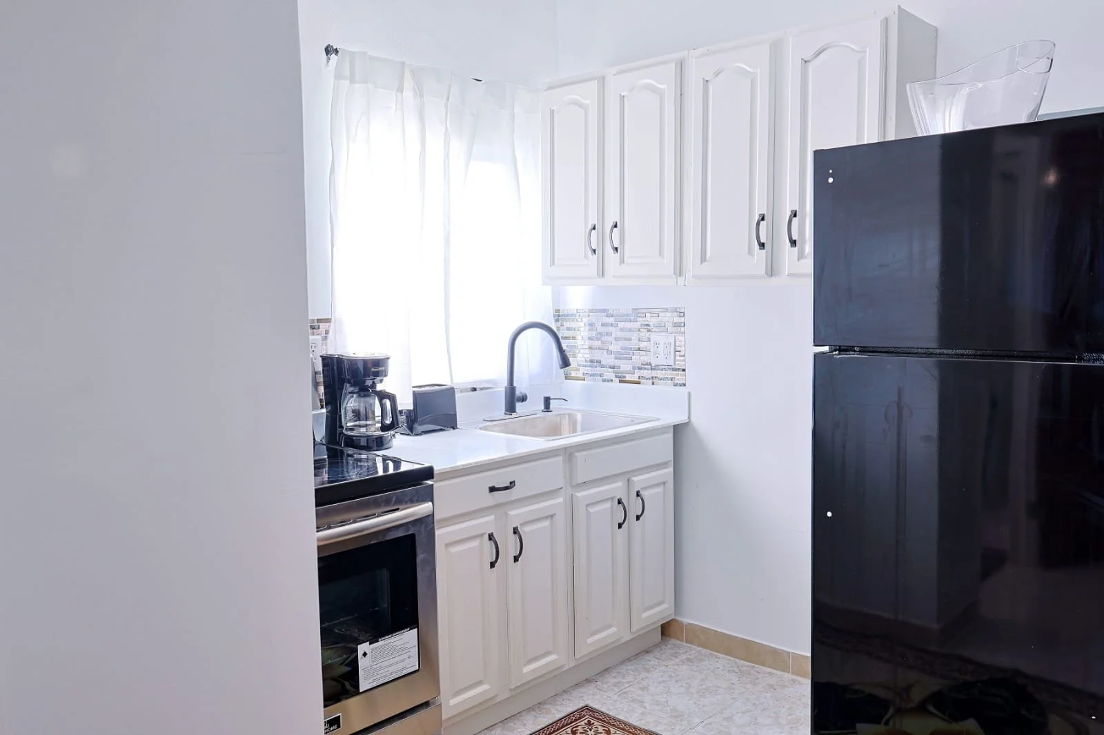 Small kitchen with white cabinets, black refrigerator, black stove, coffee maker on counter near window, and a sink with a black faucet, with a backsplash of small, multicolored tiles.