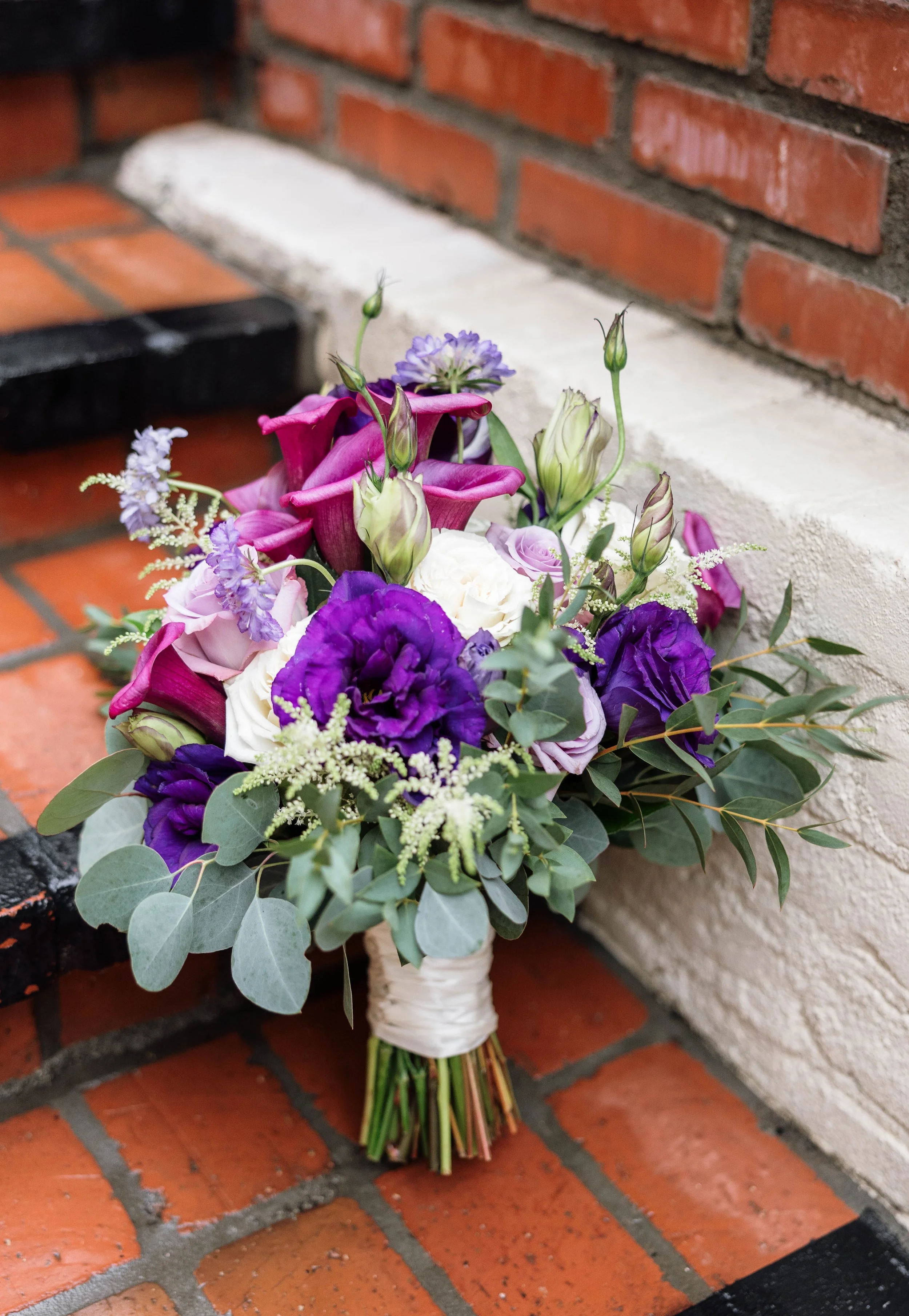 A colorful bouquet of flowers, including purple lisianthus, white roses, purple and pink calla lilies, and greenery, resting against a brick and white stone wall.
