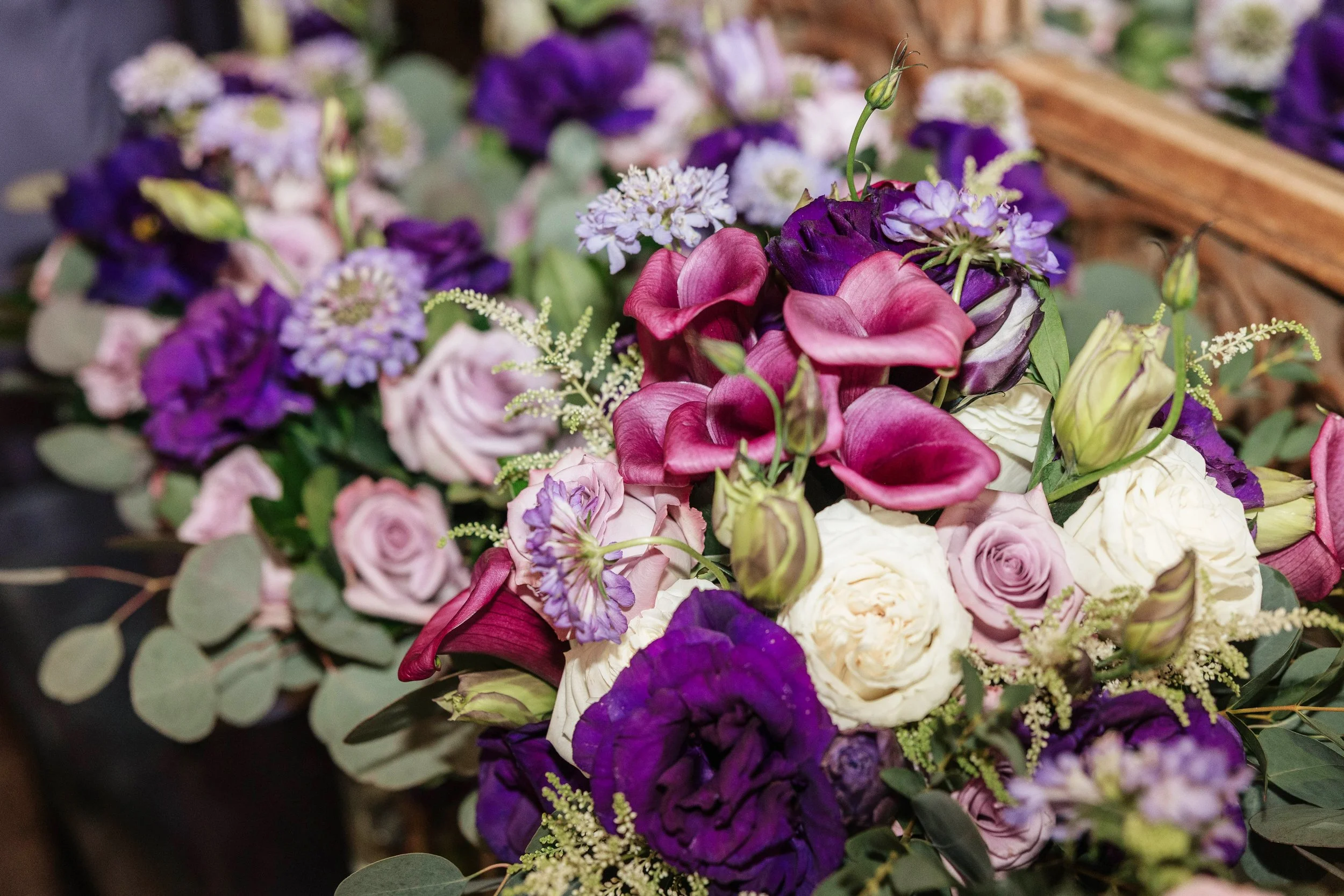 A bouquet of various flowers including purple, pink, and white roses, calla lilies, and other small purple and white flowers, surrounded by green leaves.