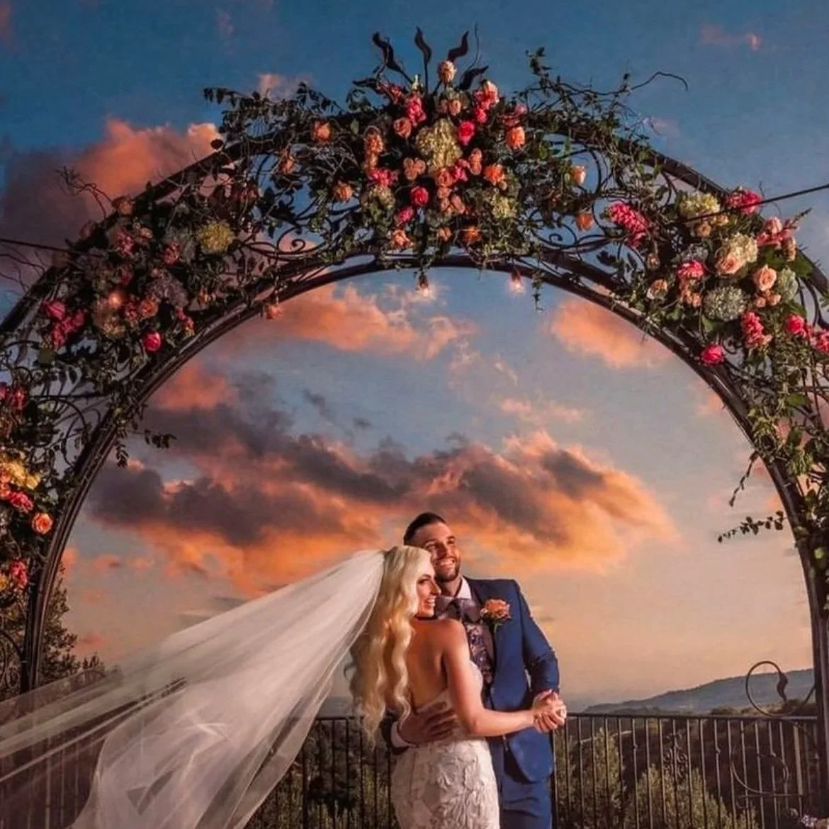 A newlywed couple stands under a floral wedding arch at sunset, with mountains in the background.