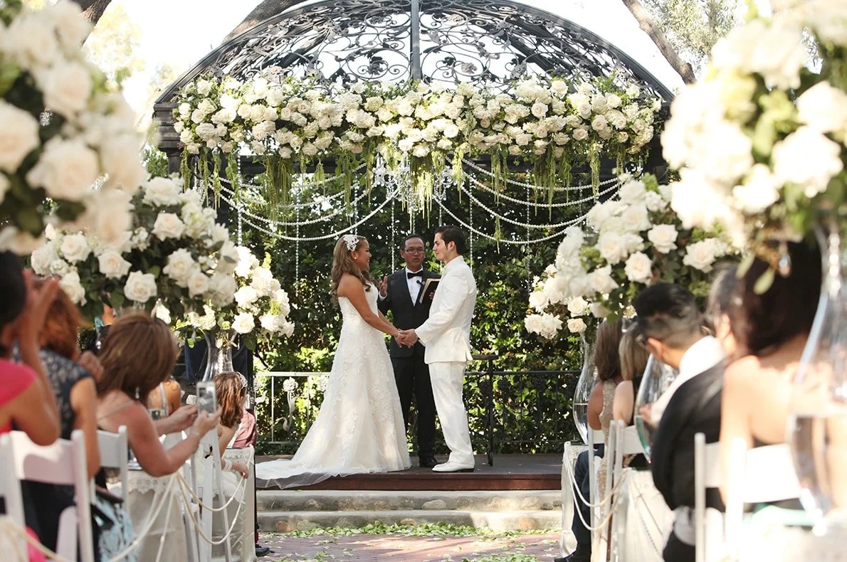A wedding ceremony taking place outdoors under a decorated arch with white flowers and hanging string lights. The bride and groom are holding hands, exchanging vows, with an officiant standing behind them. Guests are seated on either side, some taking photos, during daytime.