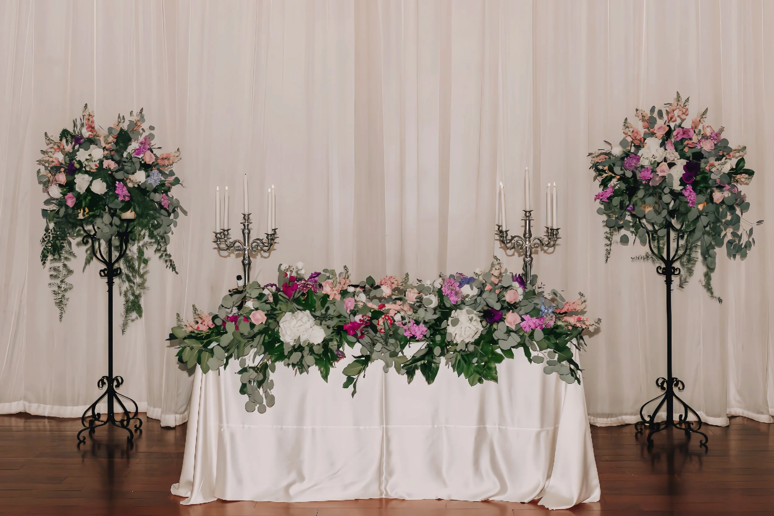 Elegant wedding reception table with white tablecloth, floral centerpieces, and tall silver candelabras against a cream-colored curtain backdrop.