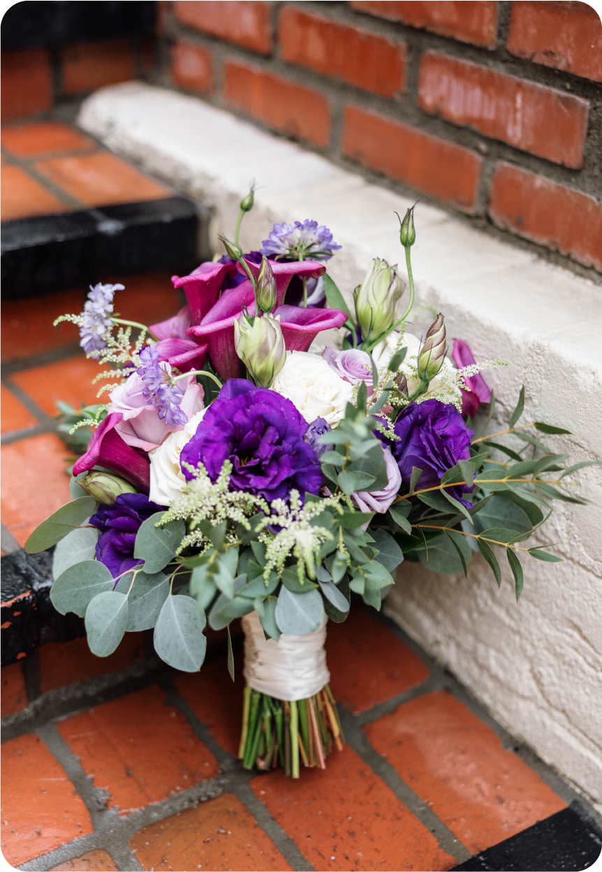 A colorful bouquet of flowers, including purple, white, and pink blossoms, with green foliage and buds, resting on a brick-and-white wall surface.
