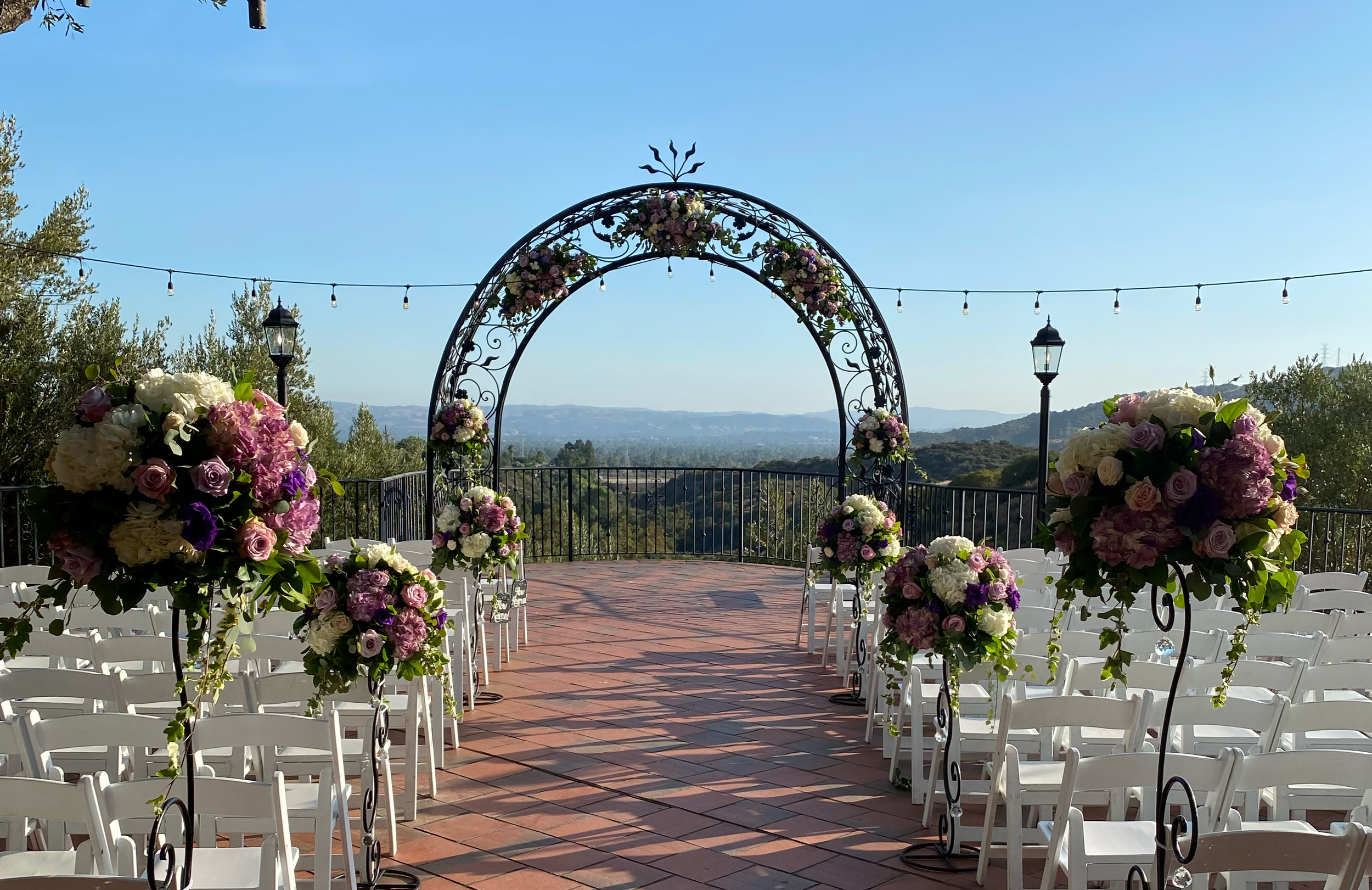 Outdoor wedding ceremony setup with white chairs, pink and purple flower arrangements, and a decorative wrought iron arch on a brick patio overlooking a scenic landscape with trees and hills.
