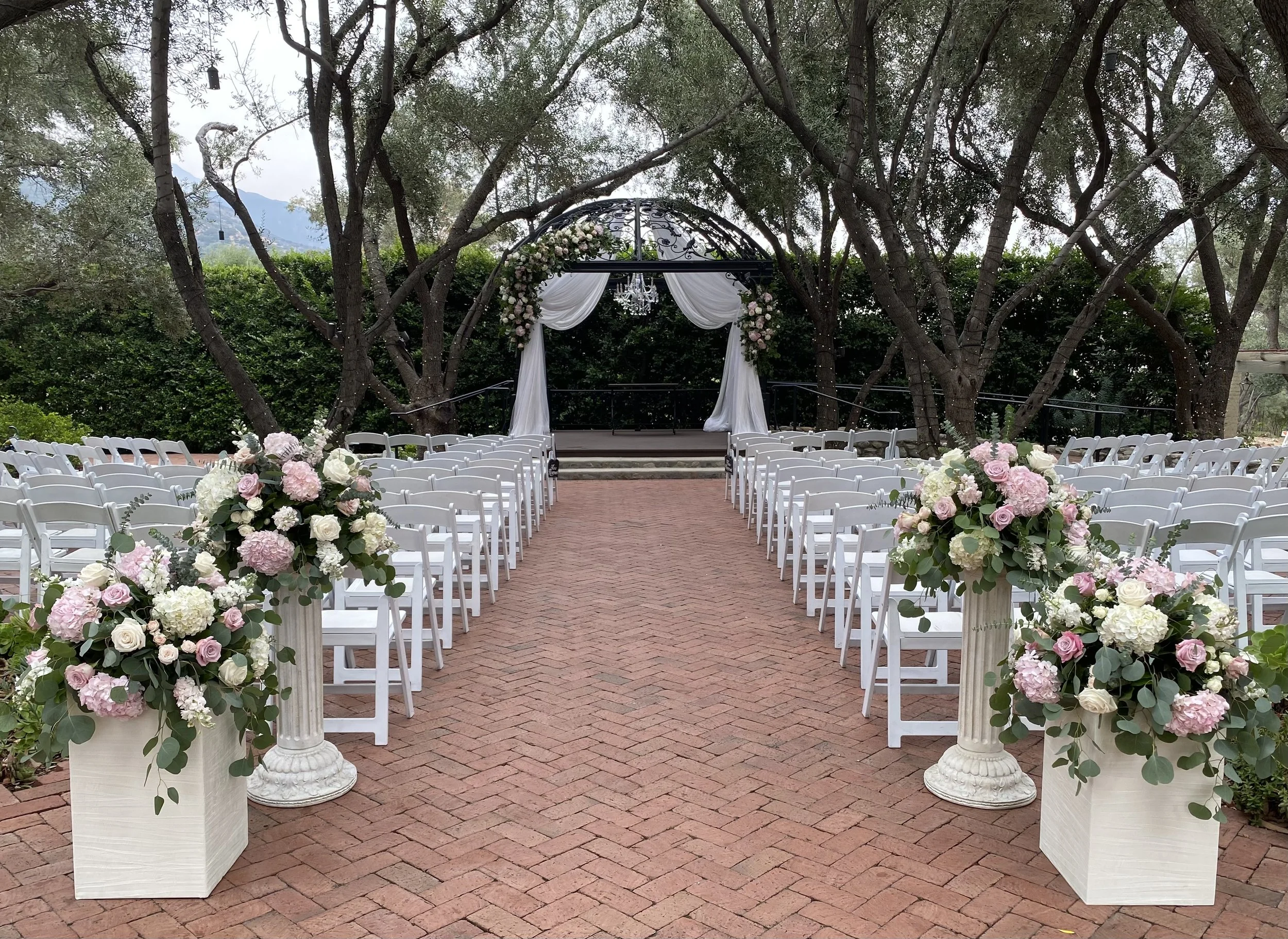 Outdoor wedding ceremony setup with white chairs, floral arrangements, and an arch decorated with draped white fabric and flowers, set on a brick pathway under trees.