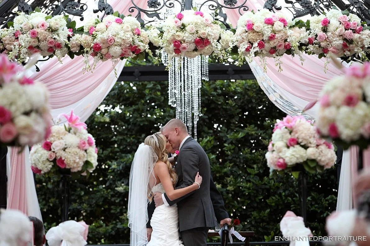 A bride and groom kiss under a floral wedding arch decorated with pink and white roses, hydrangeas, pink drapery, and crystal chandeliers during an outdoor wedding ceremony.