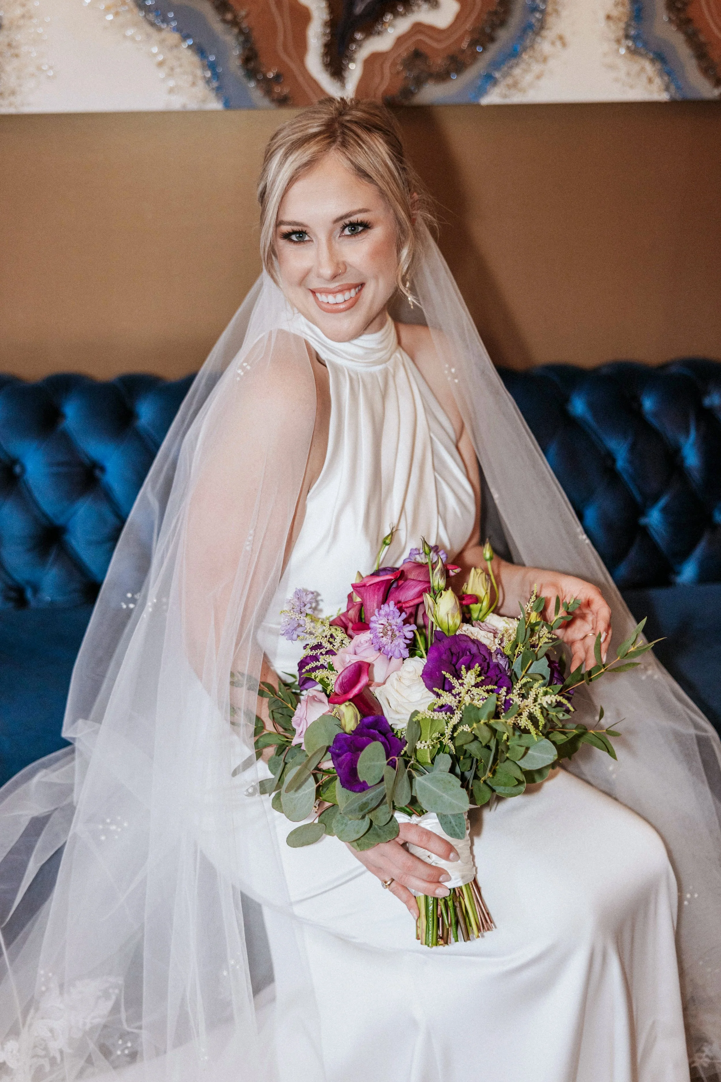 Bride in a white wedding dress with veil sitting on a dark blue tufted sofa, smiling, holding a colorful bouquet of pink, purple, and white flowers.