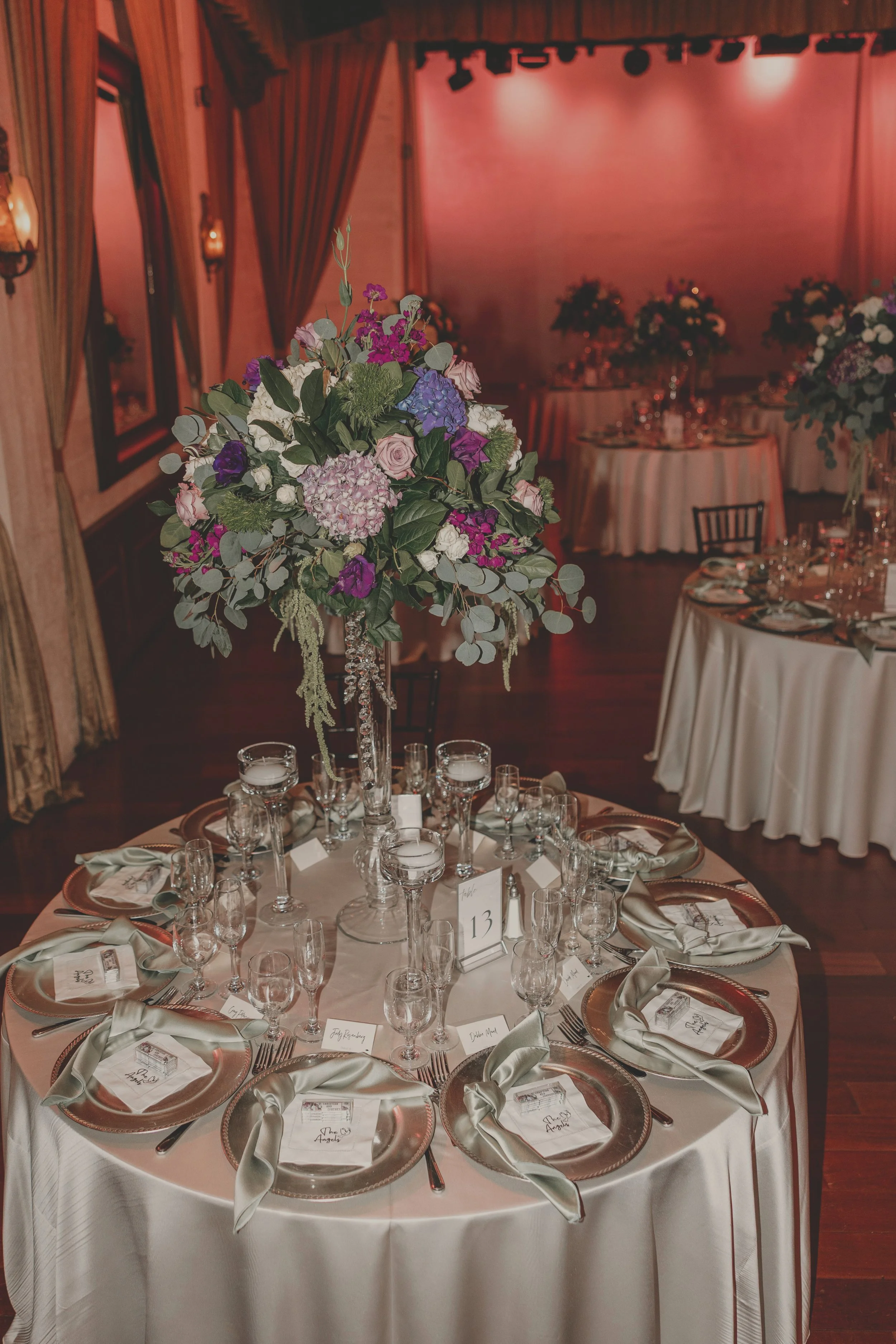 A decorated banquet table at a wedding reception with a tall floral centerpiece, place settings with plates, napkins, and glasses, and a table number, all set in a warmly lit event hall.