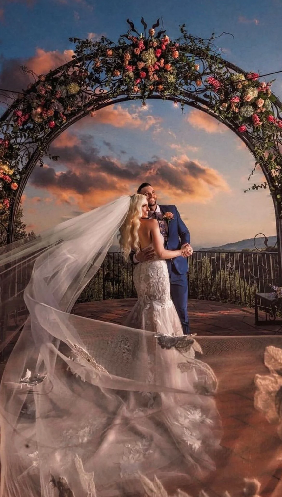 A bride and groom dancing on a decorated outdoor terrace at sunset, with an arch of flowers and sky filled with colorful clouds in the background.