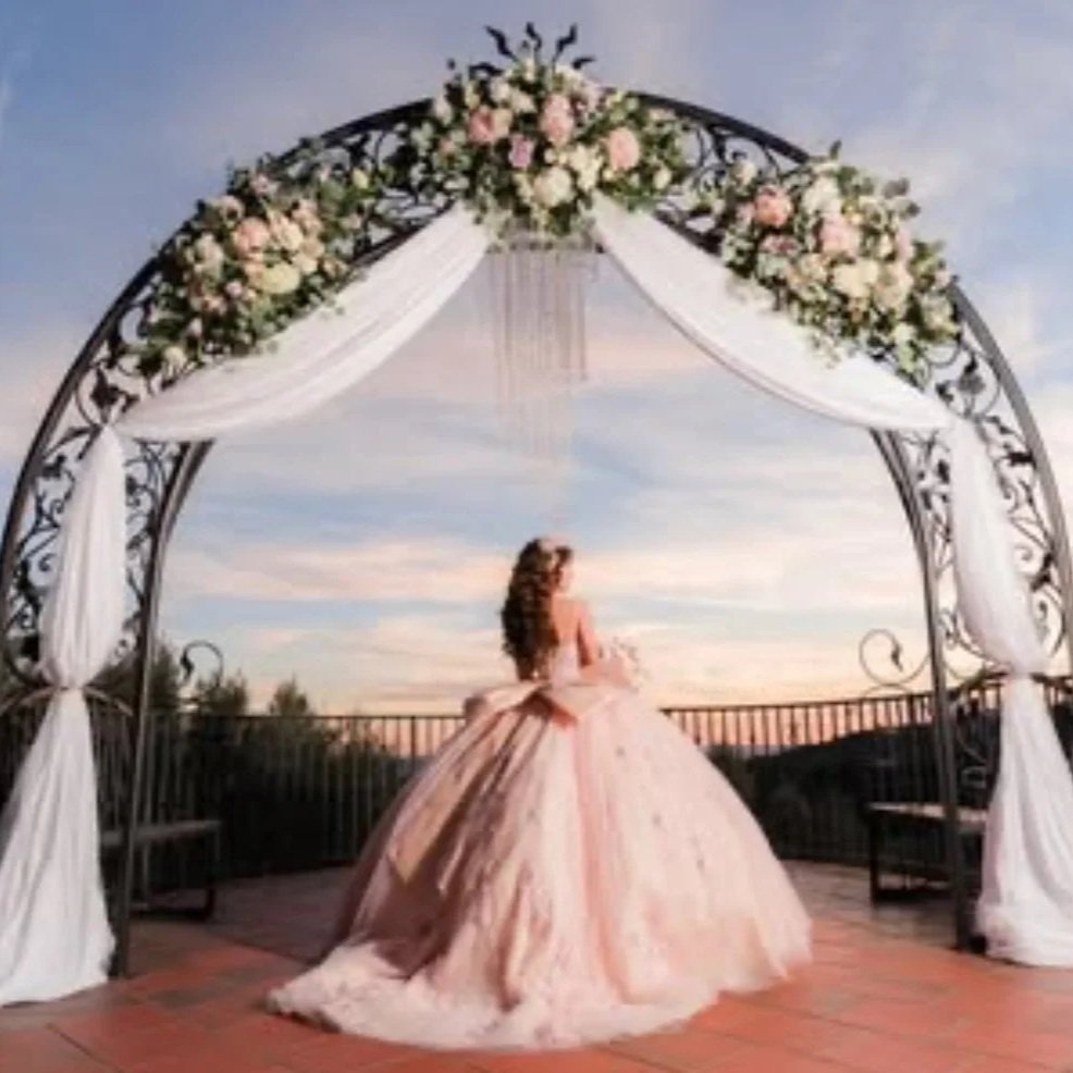 A woman in a pink ball gown standing under a decorative arch decorated with white and pink flowers, with draped white fabric, on a terrace during sunset.