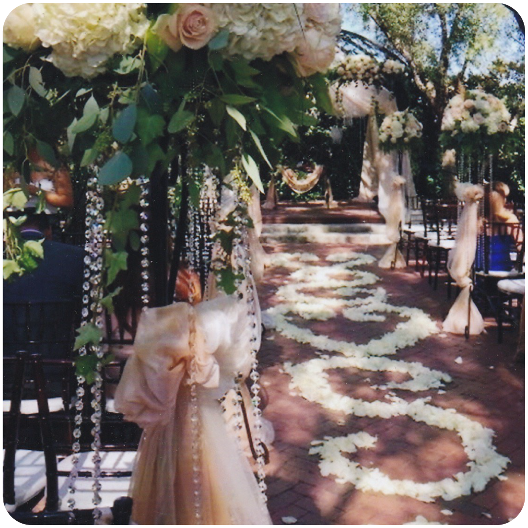 Decorated outdoor wedding aisle with floral arrangements and hanging crystals, flower petals on the ground, and chairs on either side.