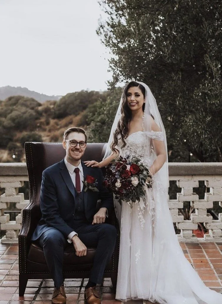 A bride and groom posed outdoors on a balcony with a scenic background of hills and trees. The groom is seated on a dark wooden chair, wearing a dark suit, white shirt, and burgundy tie, with a red boutonniere on his lapel. The bride stands next to h
