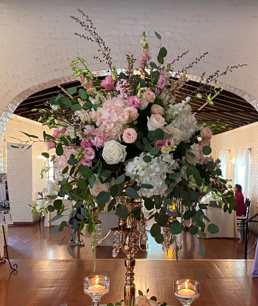 A large floral centerpiece with pink and white roses, hydrangeas, green leaves, and sprigs, placed on a golden candelabrum with hanging crystals, in a decorated event hall with candles and people in the background.