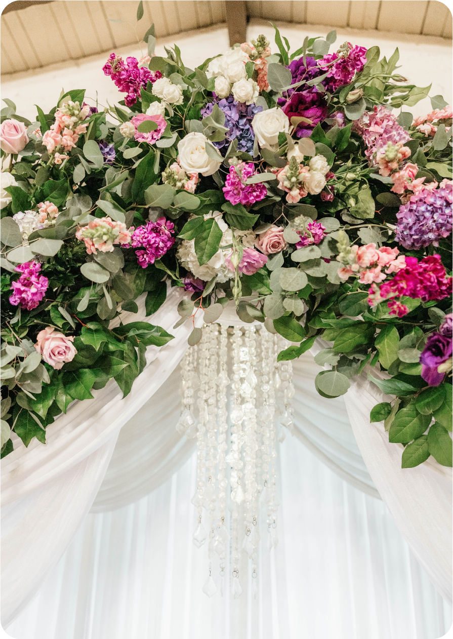 A floral arrangement with pink, purple, and white flowers, green leaves, white draped fabric, and hanging crystal decorations, likely part of wedding or event decor.