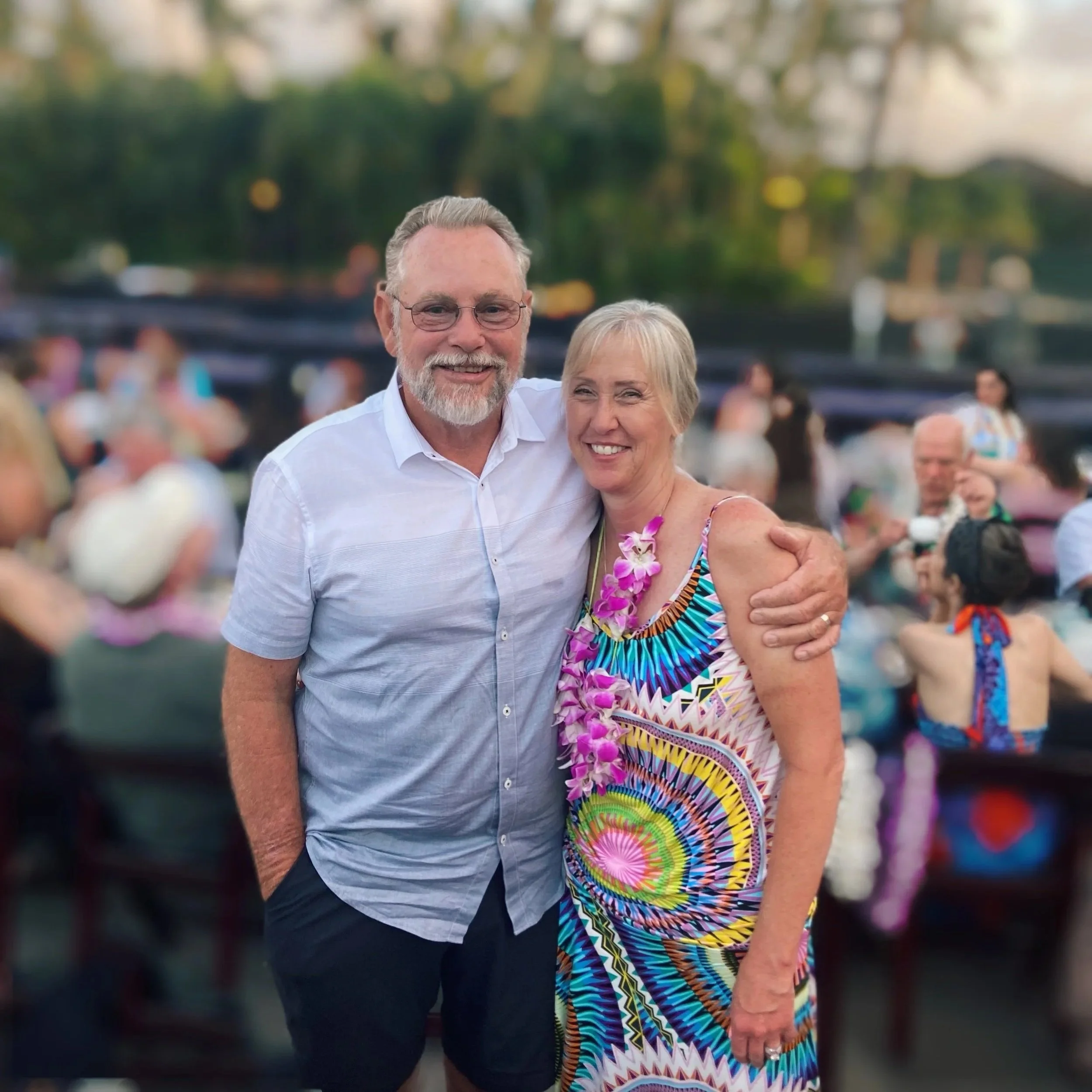 A smiling older couple at a tropical outdoor gathering, woman wearing a colorful patterned dress and purple orchid lei, man in a light blue button-up shirt, people in the background at a social event.