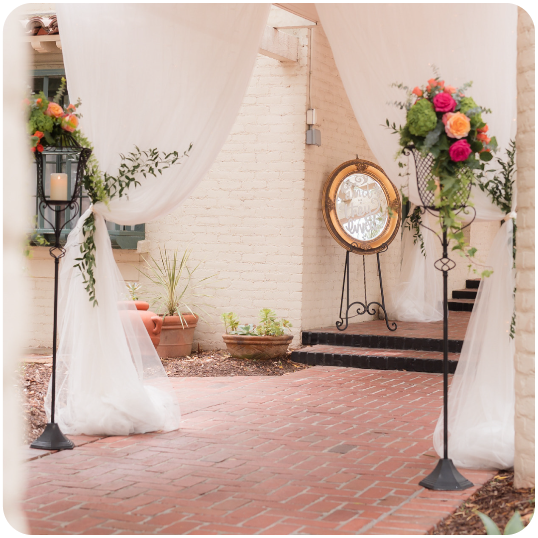 Decorative outdoor event space with white draped fabric, floral arrangements, a large mirror, and brick steps.