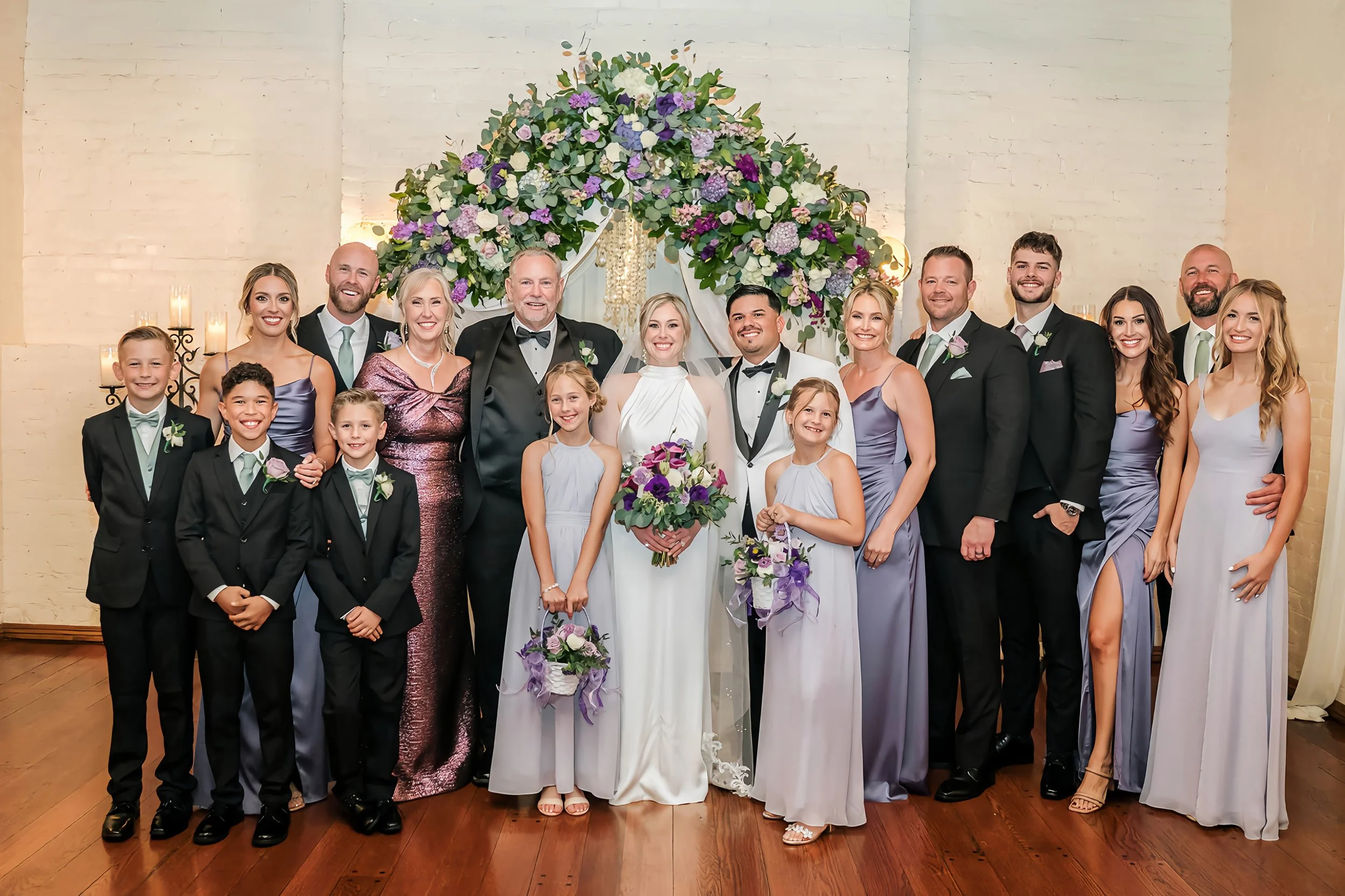 A group of wedding guests including bride, groom, children, and family members standing in front of a floral arch at a wedding celebration.