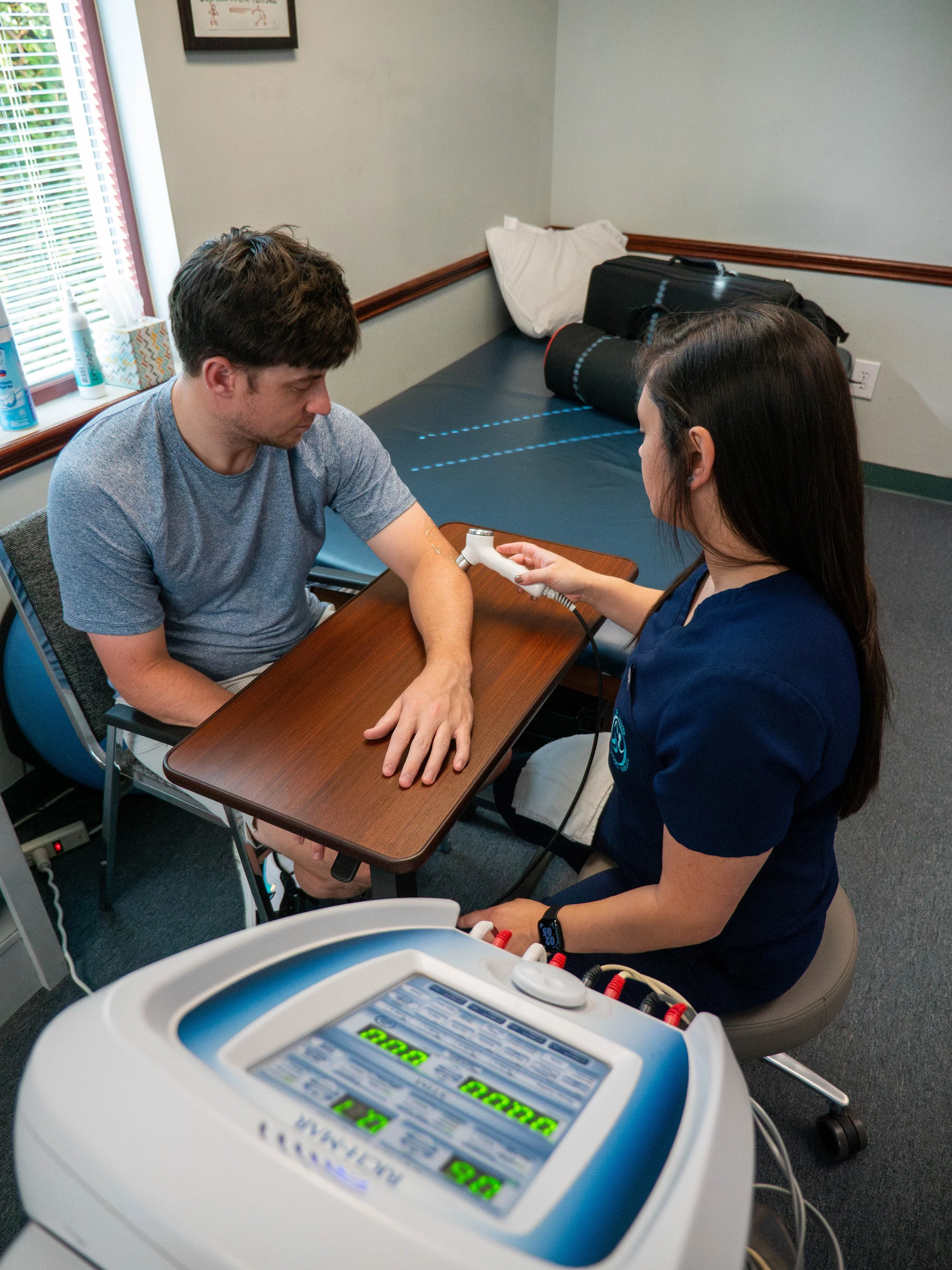 A healthcare professional performs a nerve conduction test on a man's arm in a medical office. The woman is holding a device against his forearm while he sits at a wooden table. Medical equipment and a bed are visible in the background.