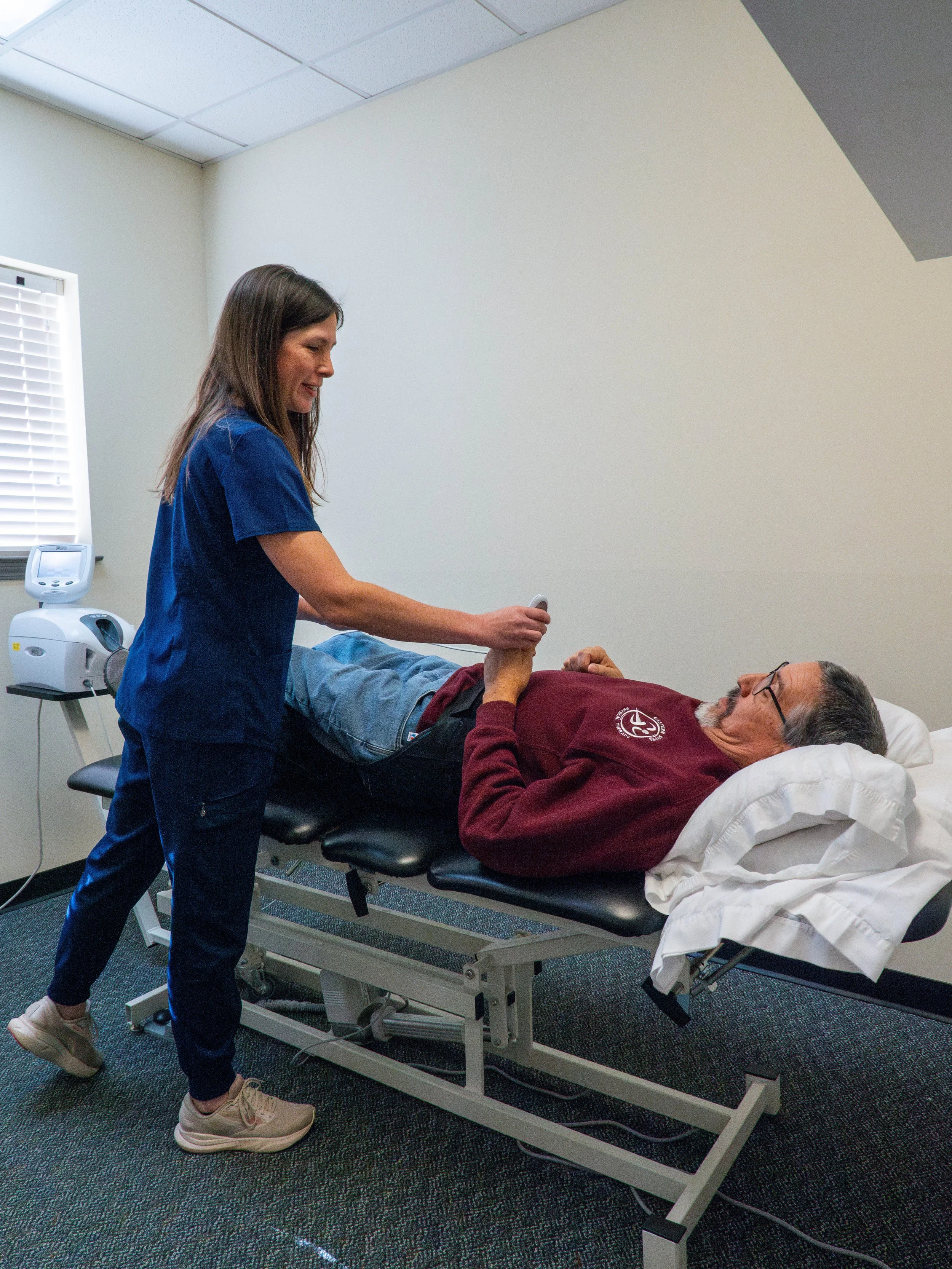 A healthcare worker in blue scrubs is holding an older man's hand while he is lying on a medical examination table, smiling.