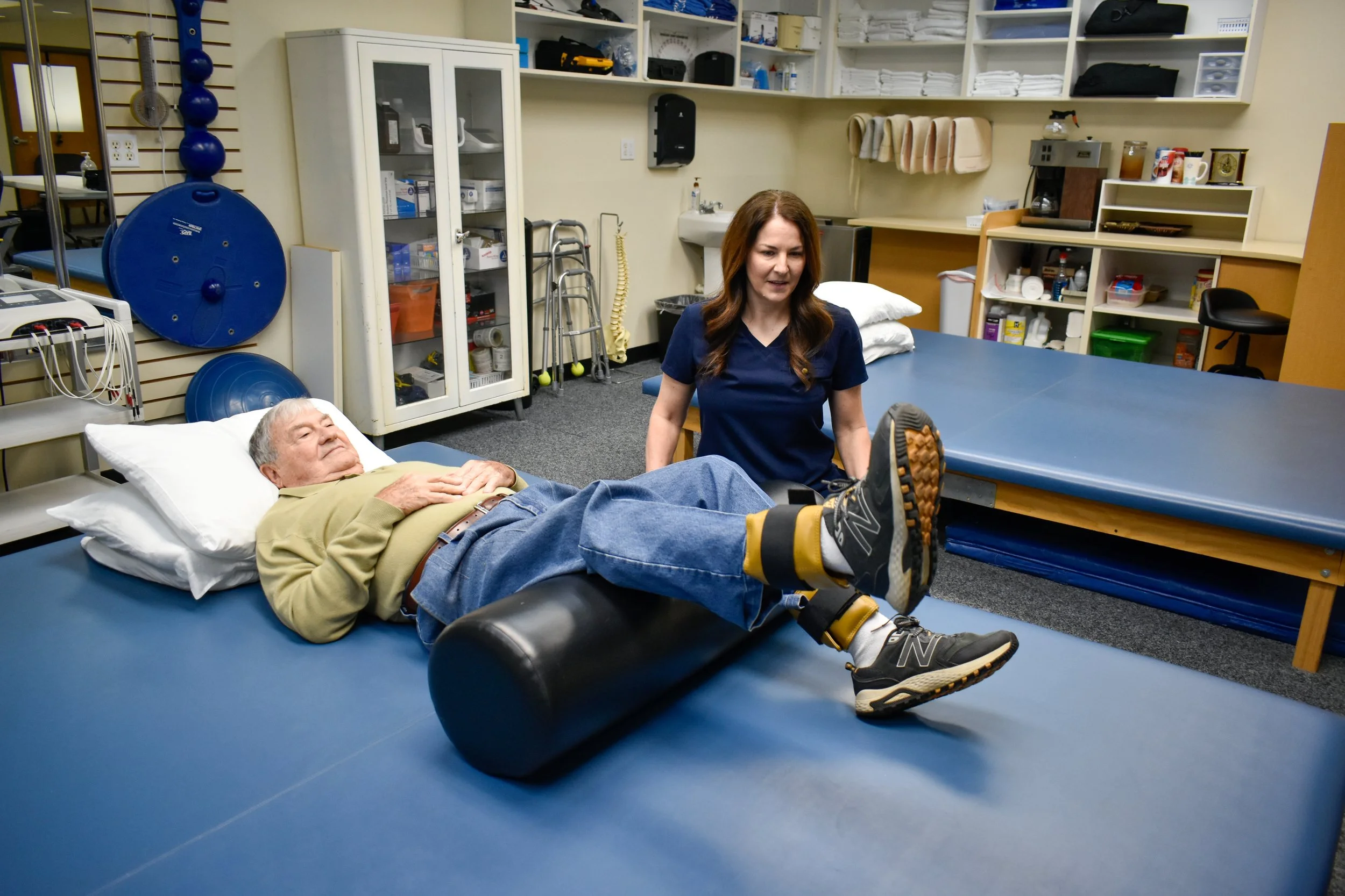 An elderly man lying on a blue therapy table with his legs elevated on a foam roller while a female therapist in dark scrubs assists him during a physiotherapy session in a medical facility.