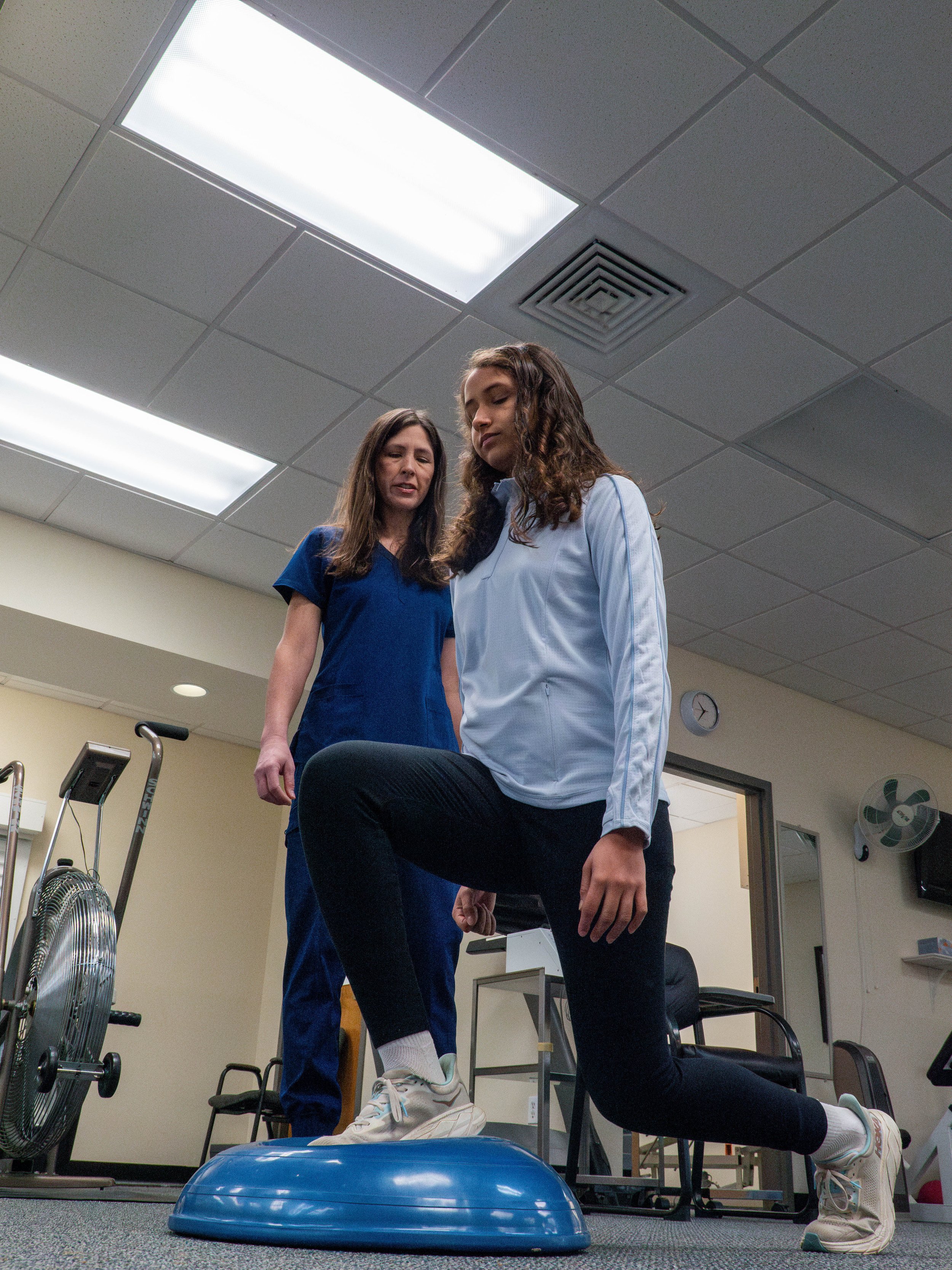 A woman in white shirt performing a balance exercise on a blue disc with a trainer in blue scrubs supervising.