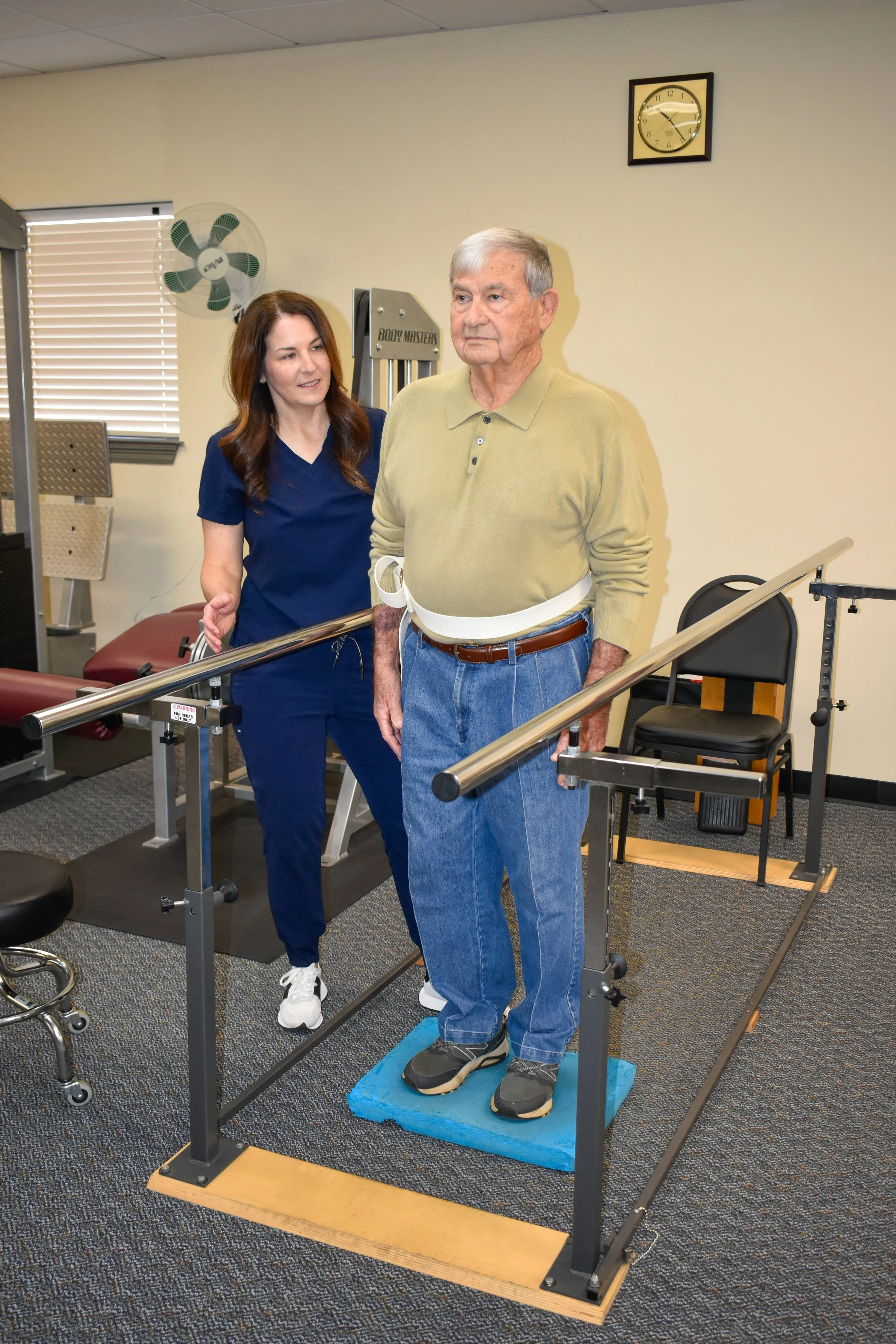Older man in a beige sweater and jeans practicing standing with support bars, guided by a female physical therapist in a navy scrubs, in a rehabilitation facility.