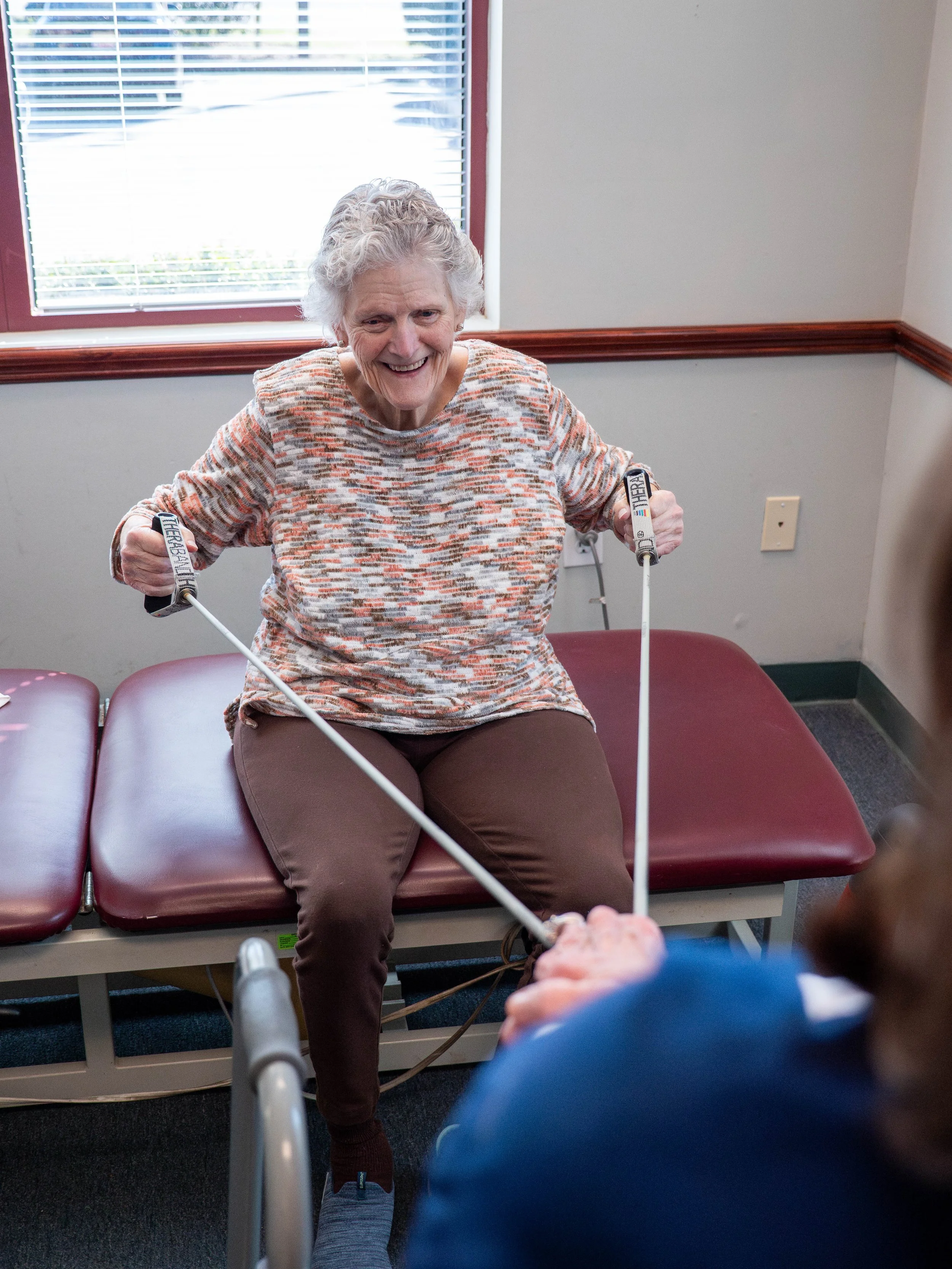 An elderly woman smiling while seated on a medical examination table, engaging in a physical therapy exercise with resistance bands.