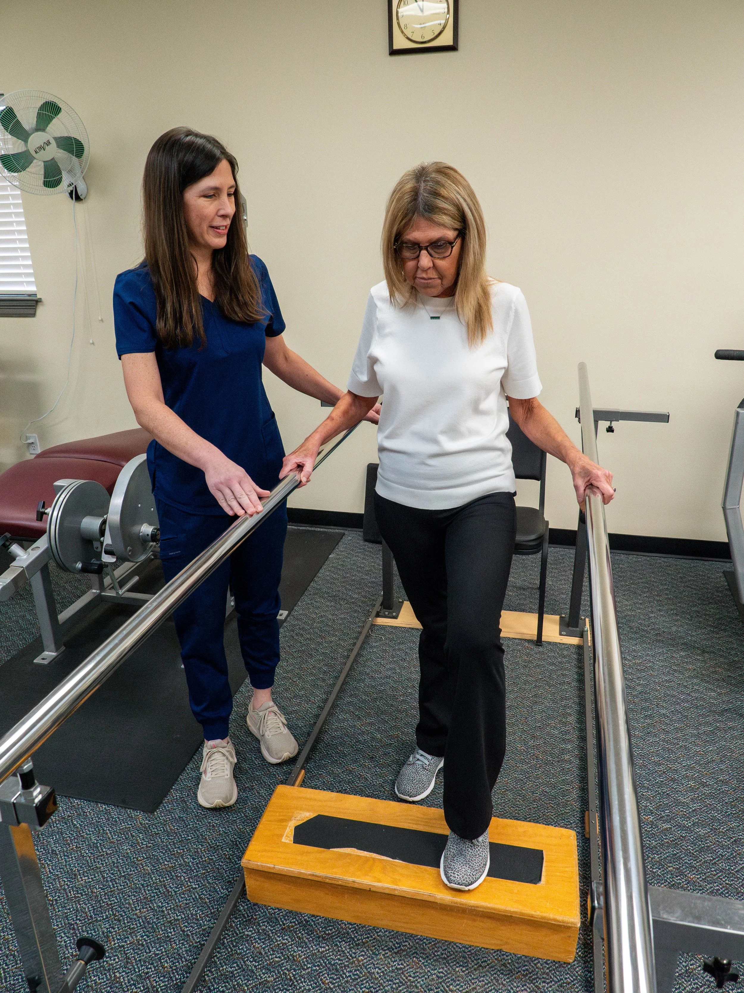 A woman in a white shirt and black pants exercises on a balance board, guided by a physical therapist in a blue uniform, in a rehabilitation clinic.